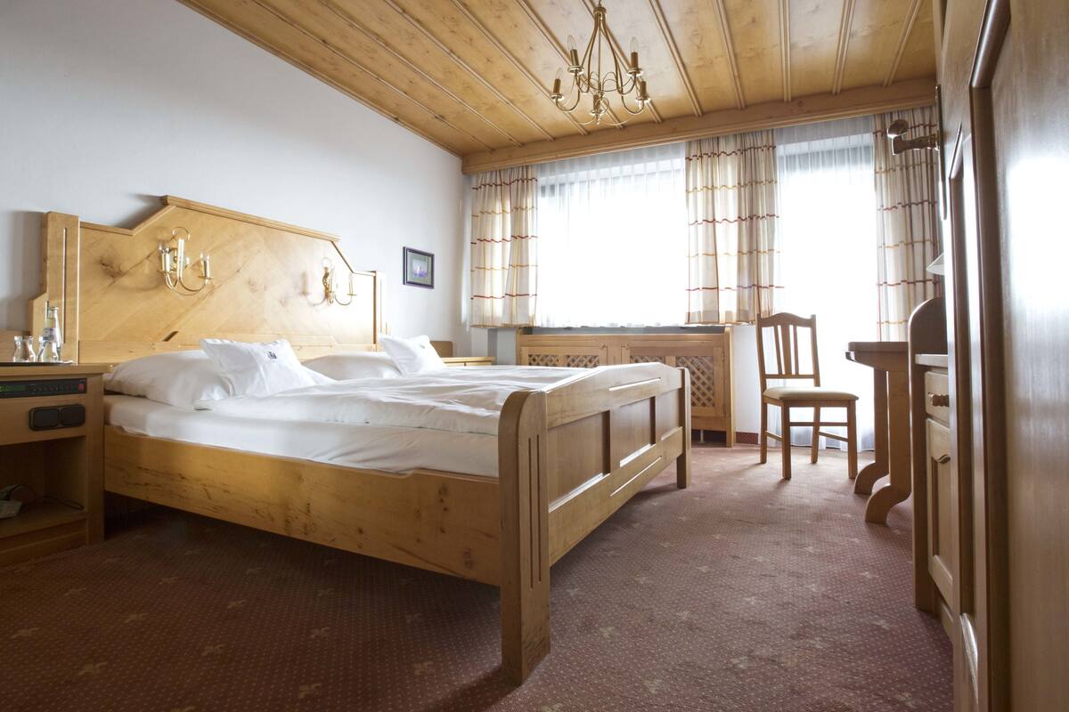 Un lit en bois dans une pièce dont le plafond est également en bois, un bureau avec une chaise et une fenêtre avec des rideaux.