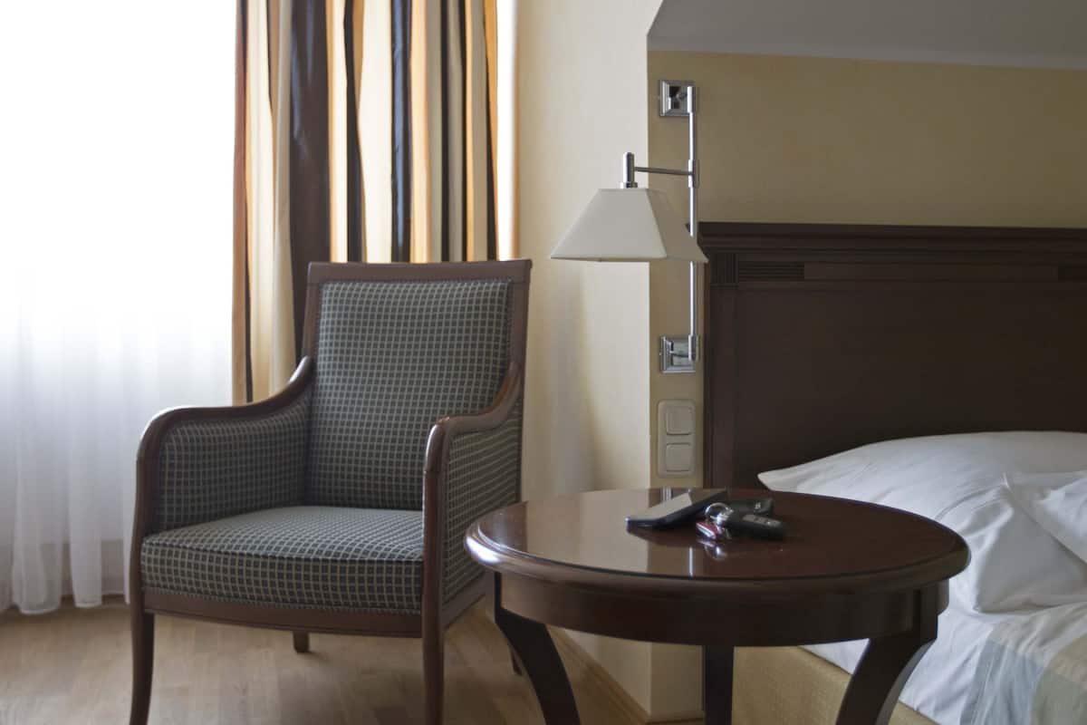 Une chambre d’hôtel avec un plancher en bois, un lit recouvert de draps blancs, une table de chevet en bois, une chaise et une lampe.