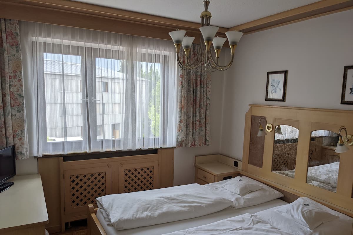 Une chambre avec deux lits, un bureau en bois, une télévision, un lustre et une fenêtre avec des rideaux.