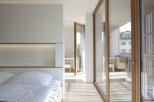 Un dormitorio moderno con una cama grande, un ventanal de vidrio que da a un balcón y vistas de edificios exteriores.
