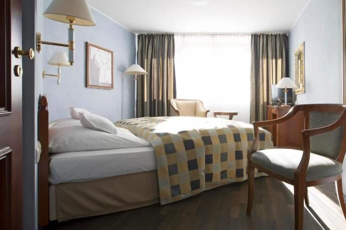 Habitación de hotel con cama, una silla, una mesa y un ventanal con cortinas.