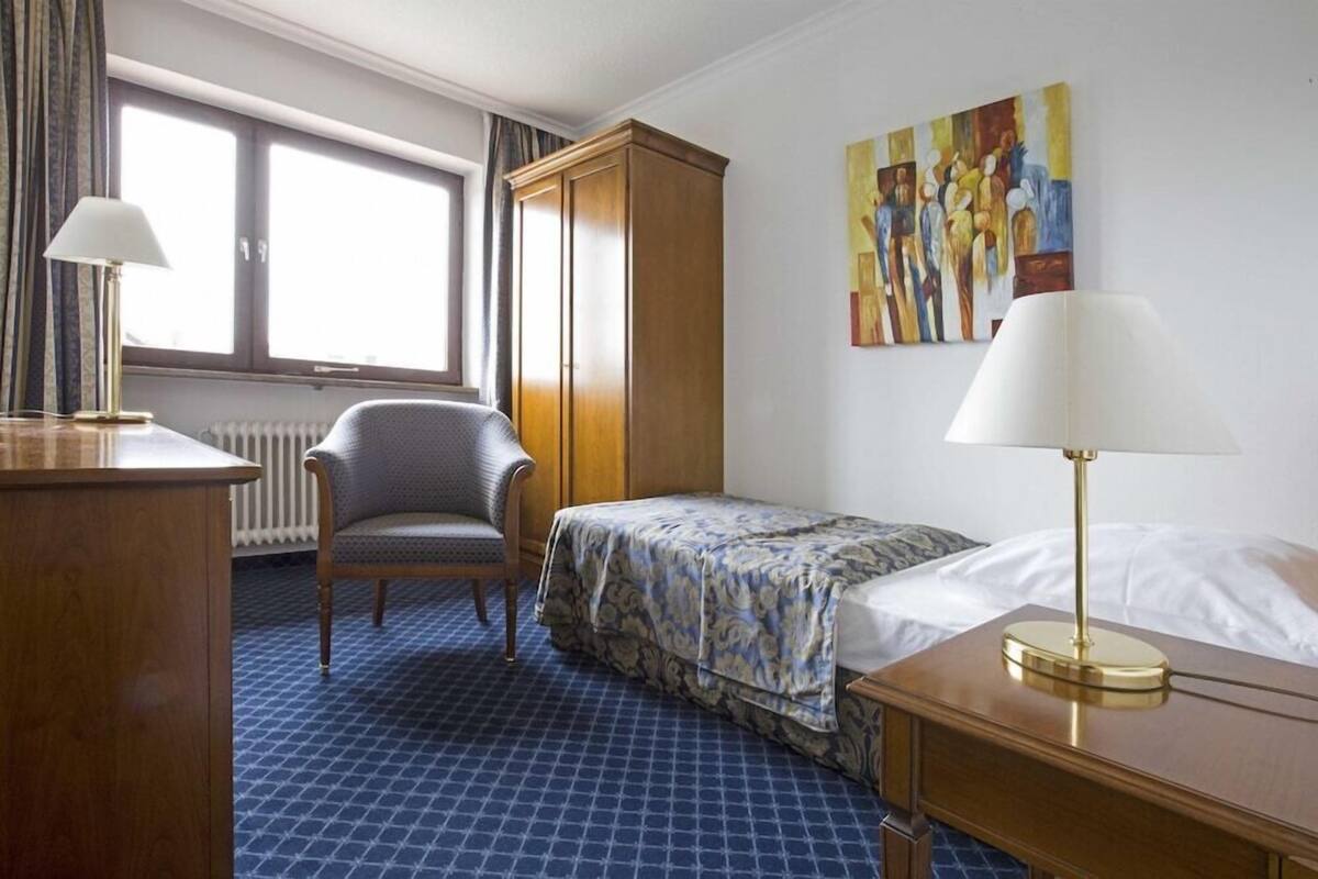 Une chambre d’hôtel avec un lit, une chaise, un bureau, un radiateur, une fenêtre et un tableau au mur.