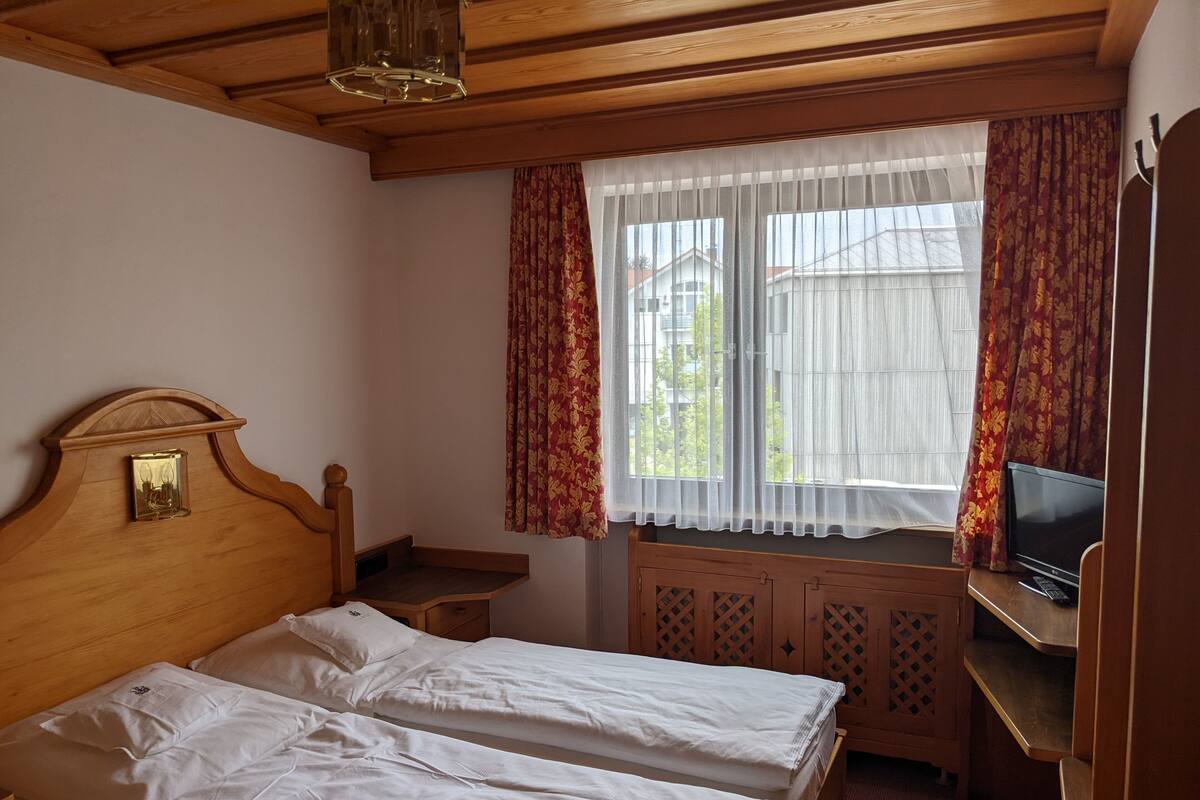 Une chambre avec deux lits, une tête de lit en bois, un téléviseur posé sur un meuble en bois et une fenêtre avec des rideaux.
