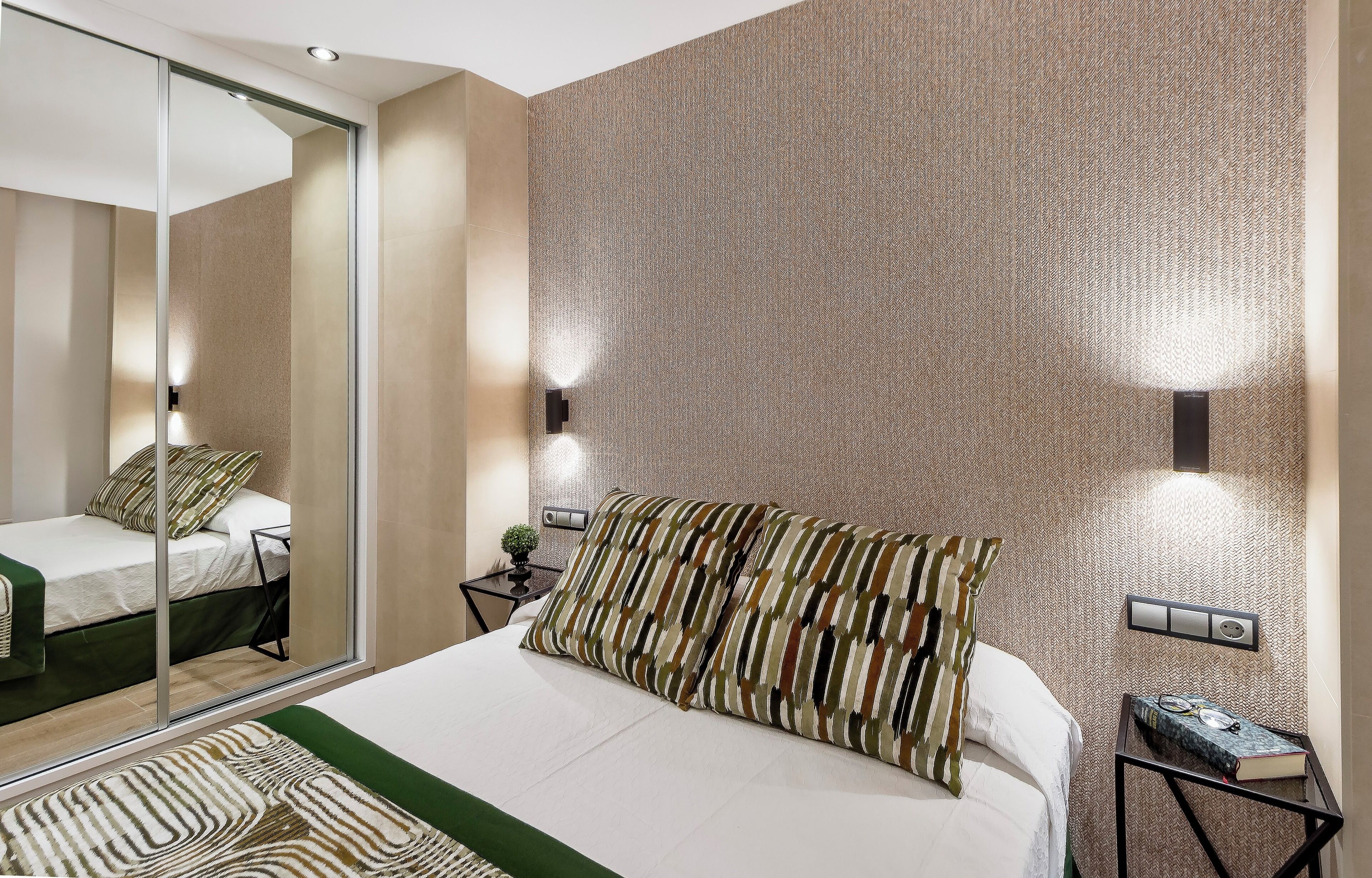 A modern hotel room with a large bed, a mirror, a nightstand, and a wall-mounted light fixture.