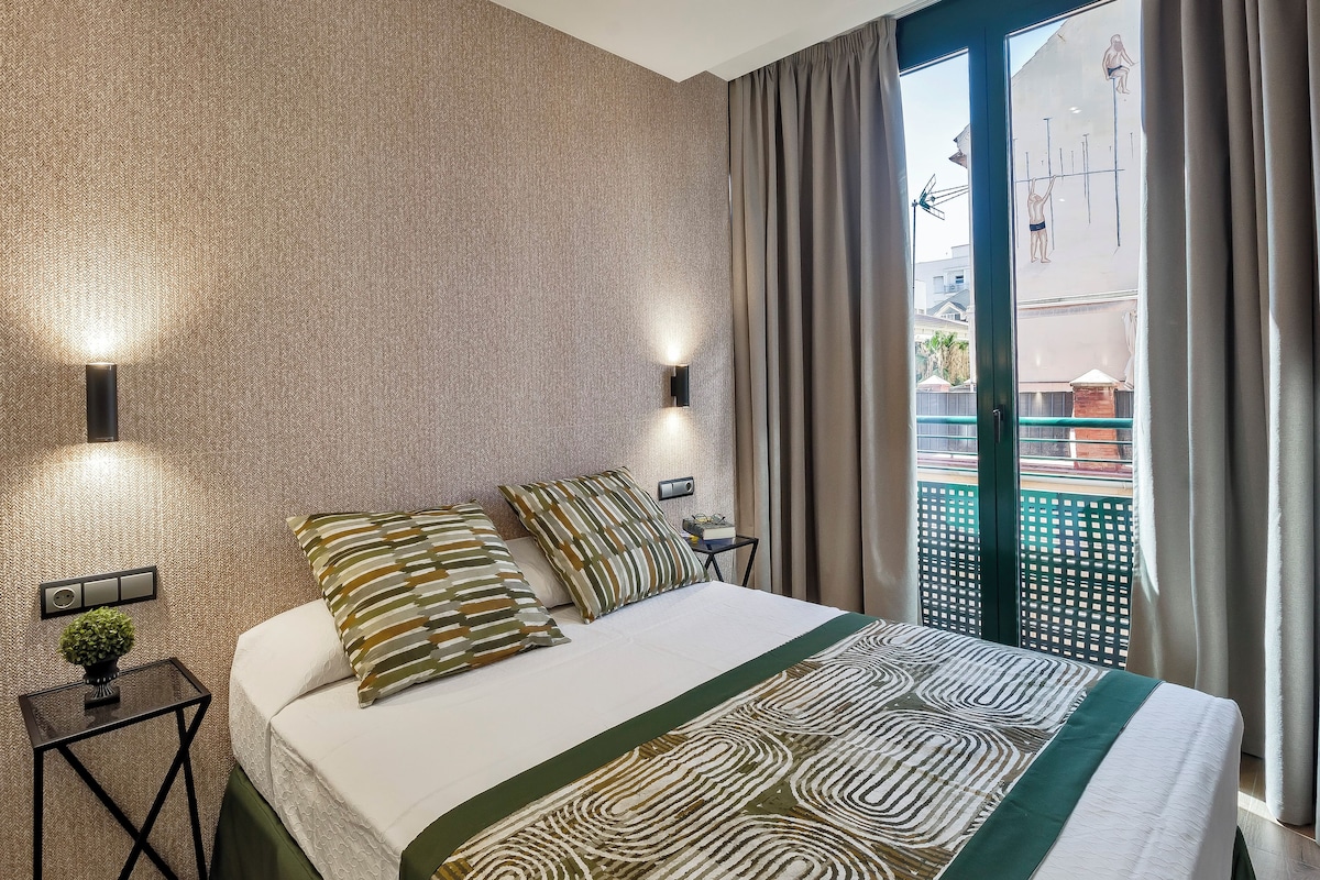 A hotel room with a large bed, two wall-mounted lights, a small table with a plant, and a view of the outside through a sliding door.