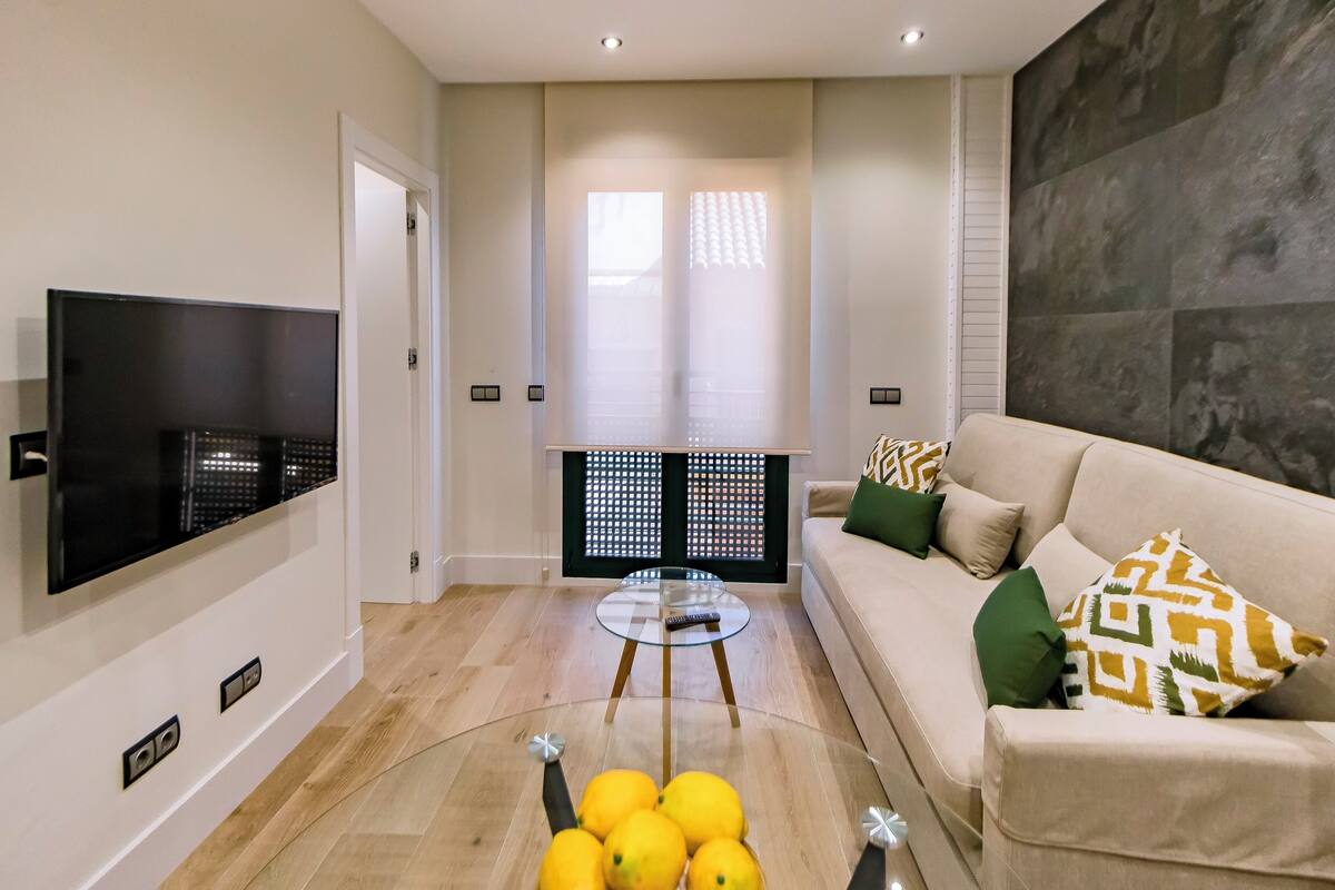 A modern living room with a flat-screen TV, a glass coffee table, a beige sofa with patterned cushions, and a wooden floor.