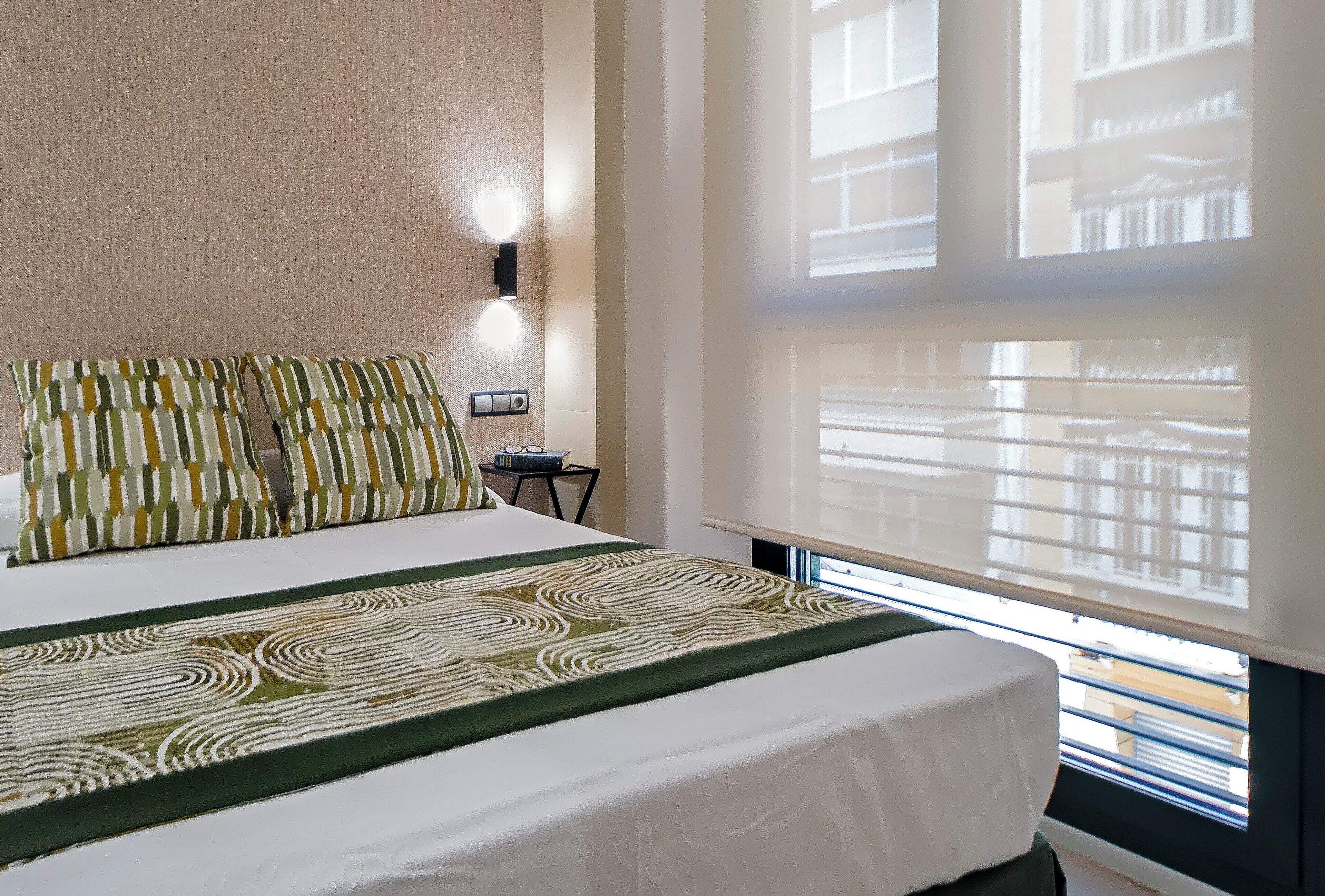Habitación de hotel con cama, luz empotrada en la pared, ventana con persianas y vista a edificios.