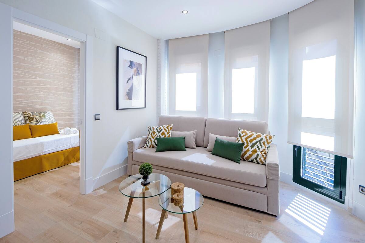 A modern living room with a sofa, a glass coffee table, and a bed with yellow bedding.