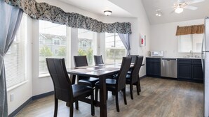 A dining area with a wooden table and chairs, a kitchen with modern appliances, and large windows with blinds. - Sunrise Inn Villas and Suites (Anacortes)