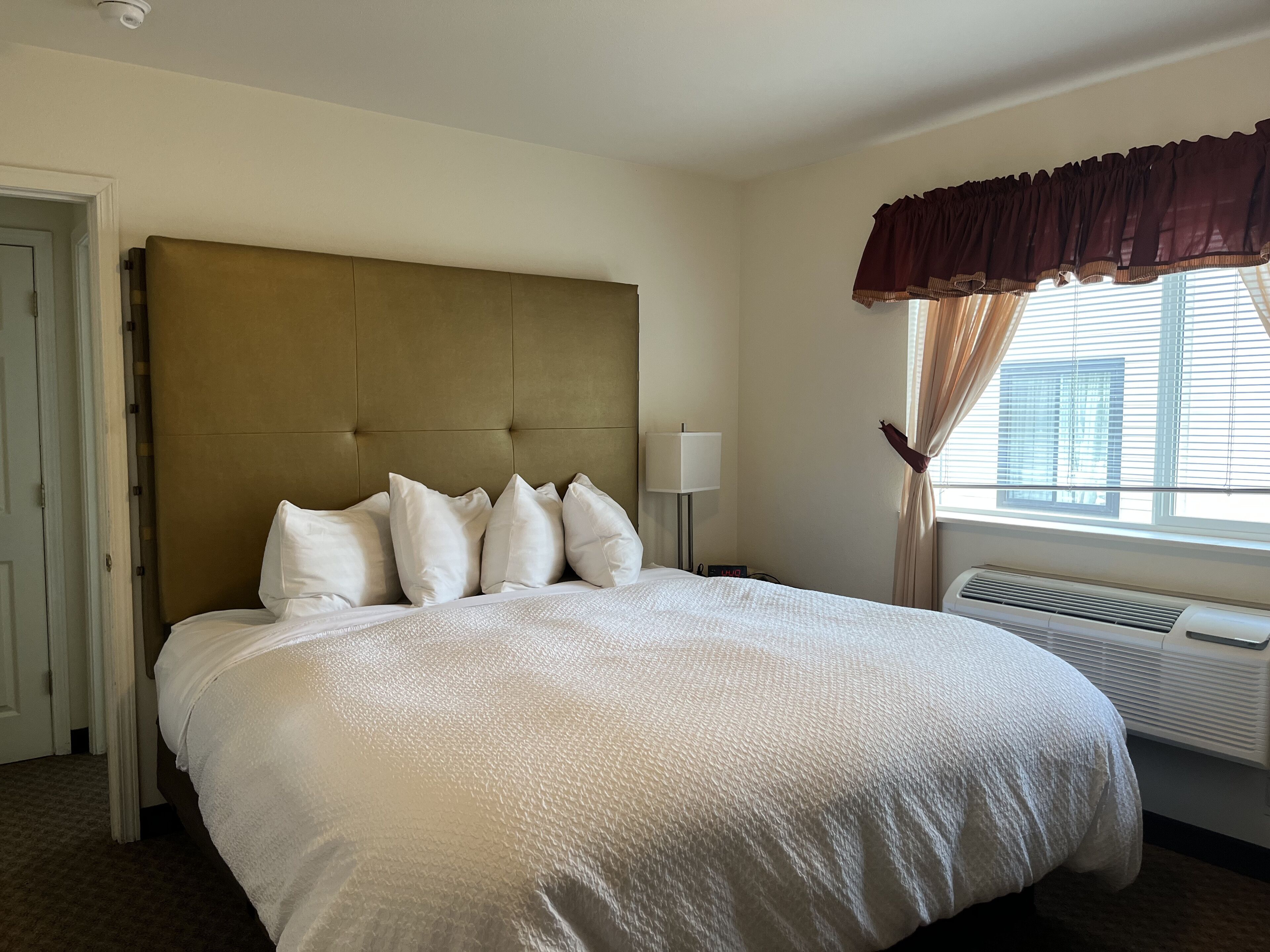 Habitación de hotel con una cama grande, cabecera, lámpara de noche, ventana con cortinas y aire acondicionado.