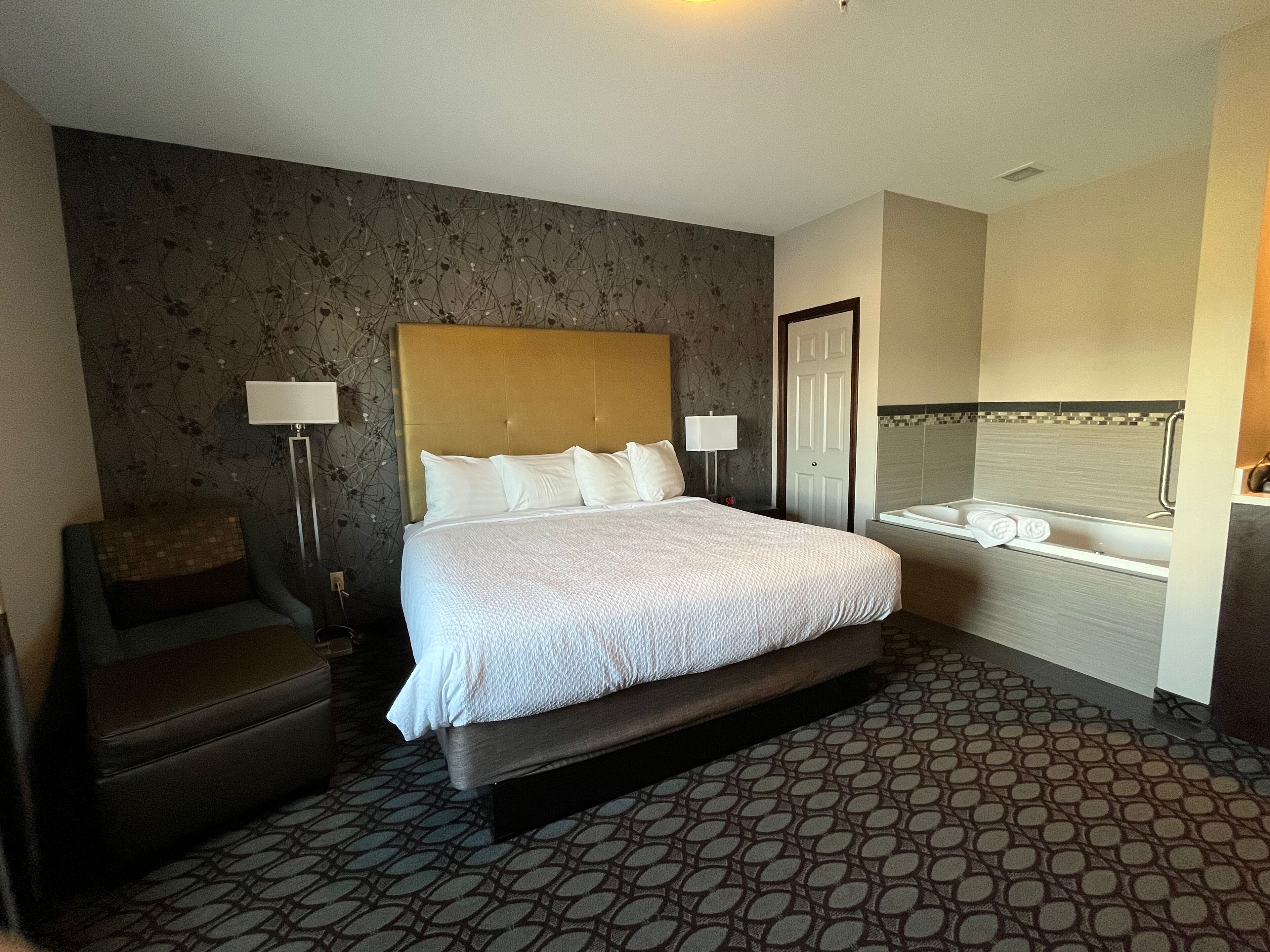 A hotel room with a large bed, a chair, a desk, a desk lamp, a bedside table, and a bathroom with a bathtub.