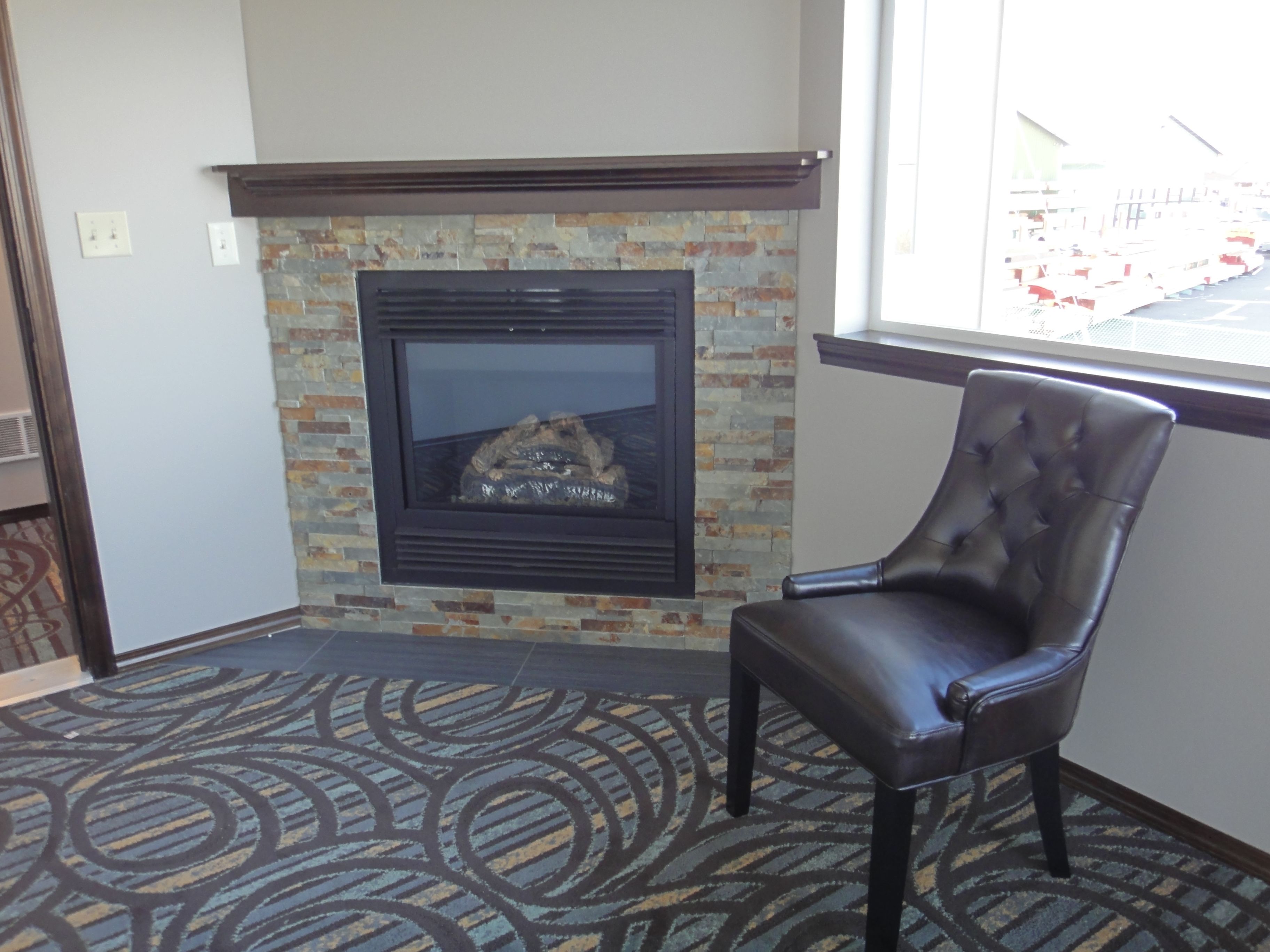 A room with a fireplace, a leather chair, patterned carpet, and a window with a view of a parking lot.
