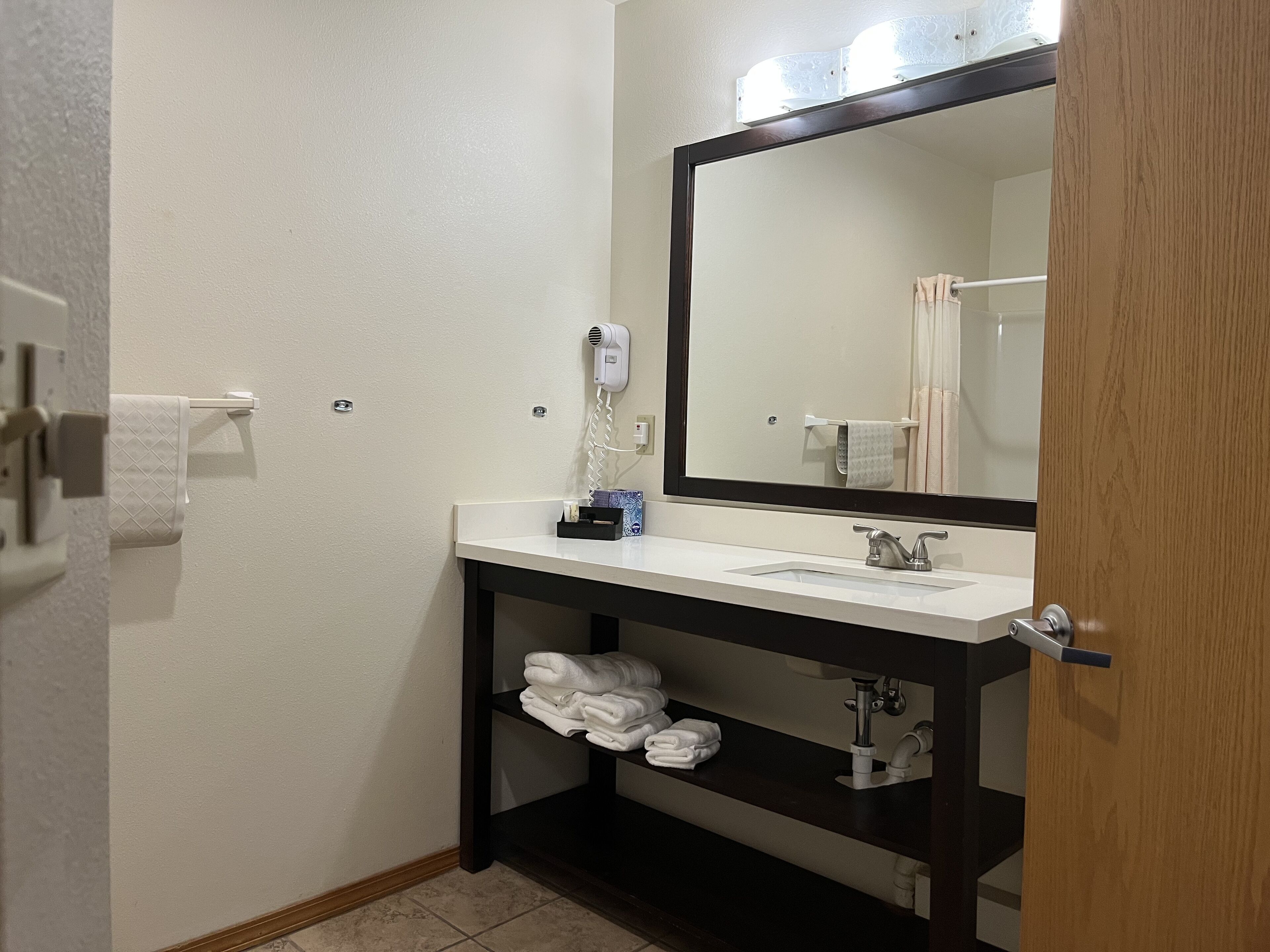 A bathroom with a white sink, a large mirror, a towel rack, a hair dryer, and a shelf with folded towels.