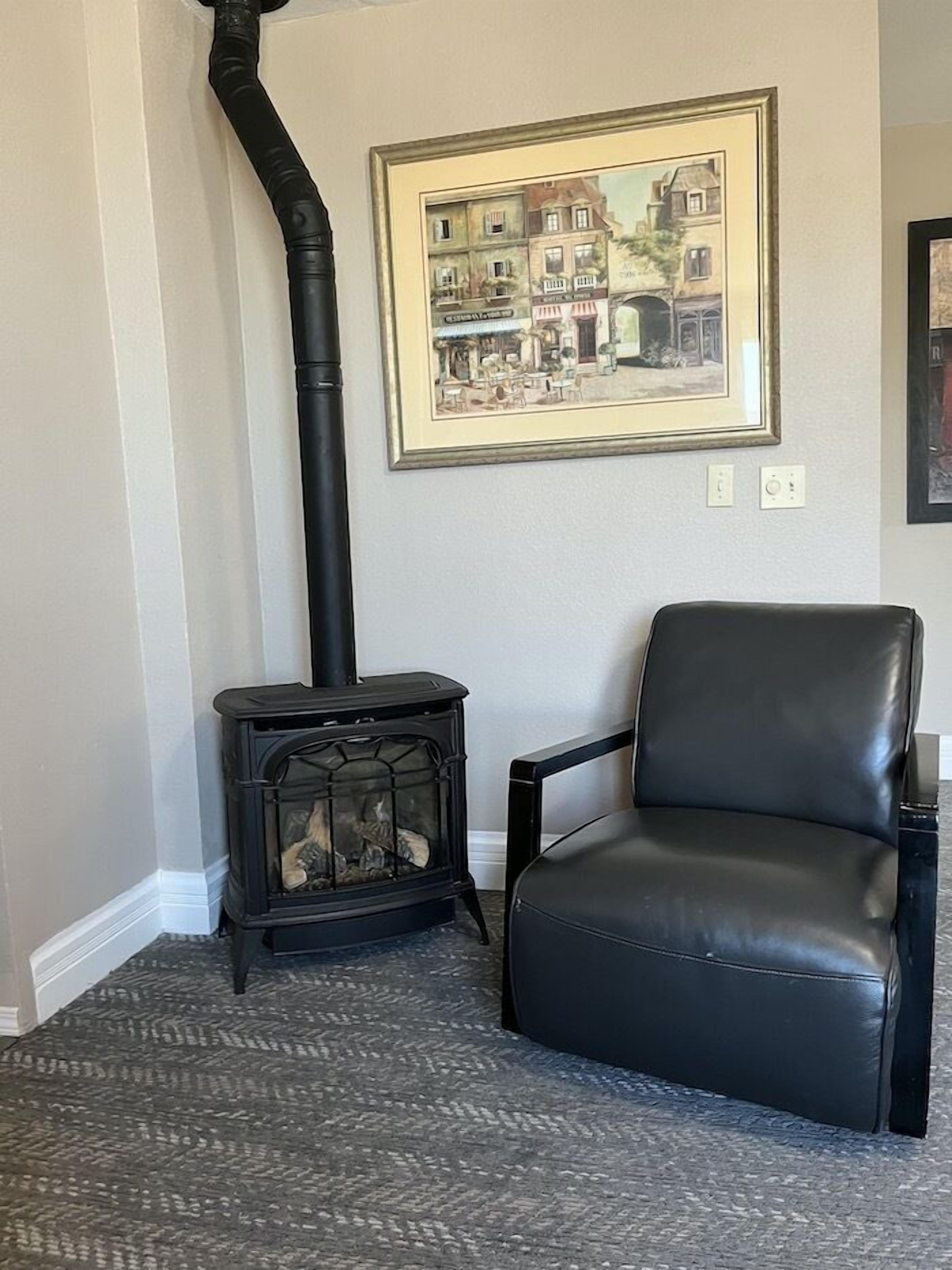 A room with a black leather chair, a black wood-burning stove, and a framed cityscape painting on the wall.