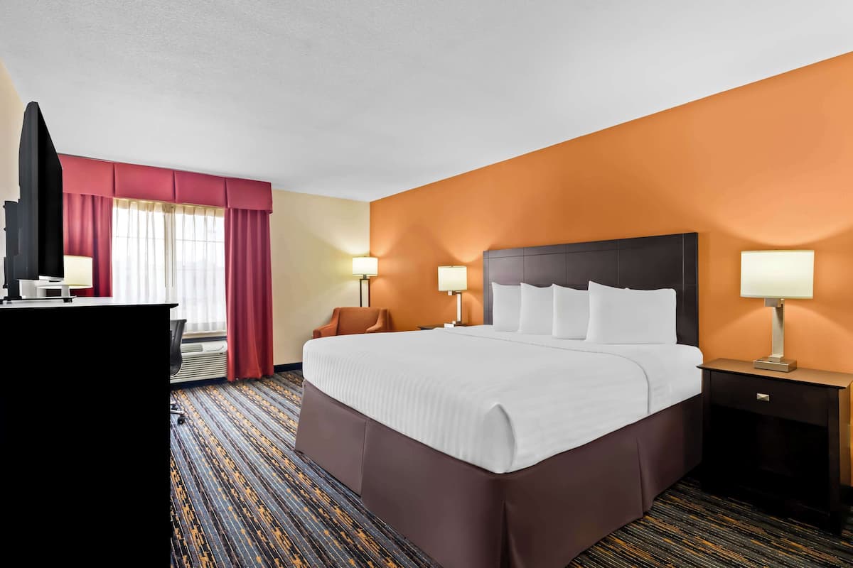 A hotel room with a large bed, two bedside tables with lamps, a desk with a TV, and a window with red curtains.