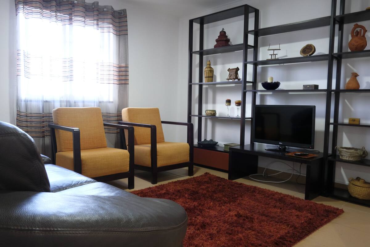 Ein Wohnzimmer mit einer Couch, einem Sessel, einem Fernseher und Regalen mit Dekoration.
