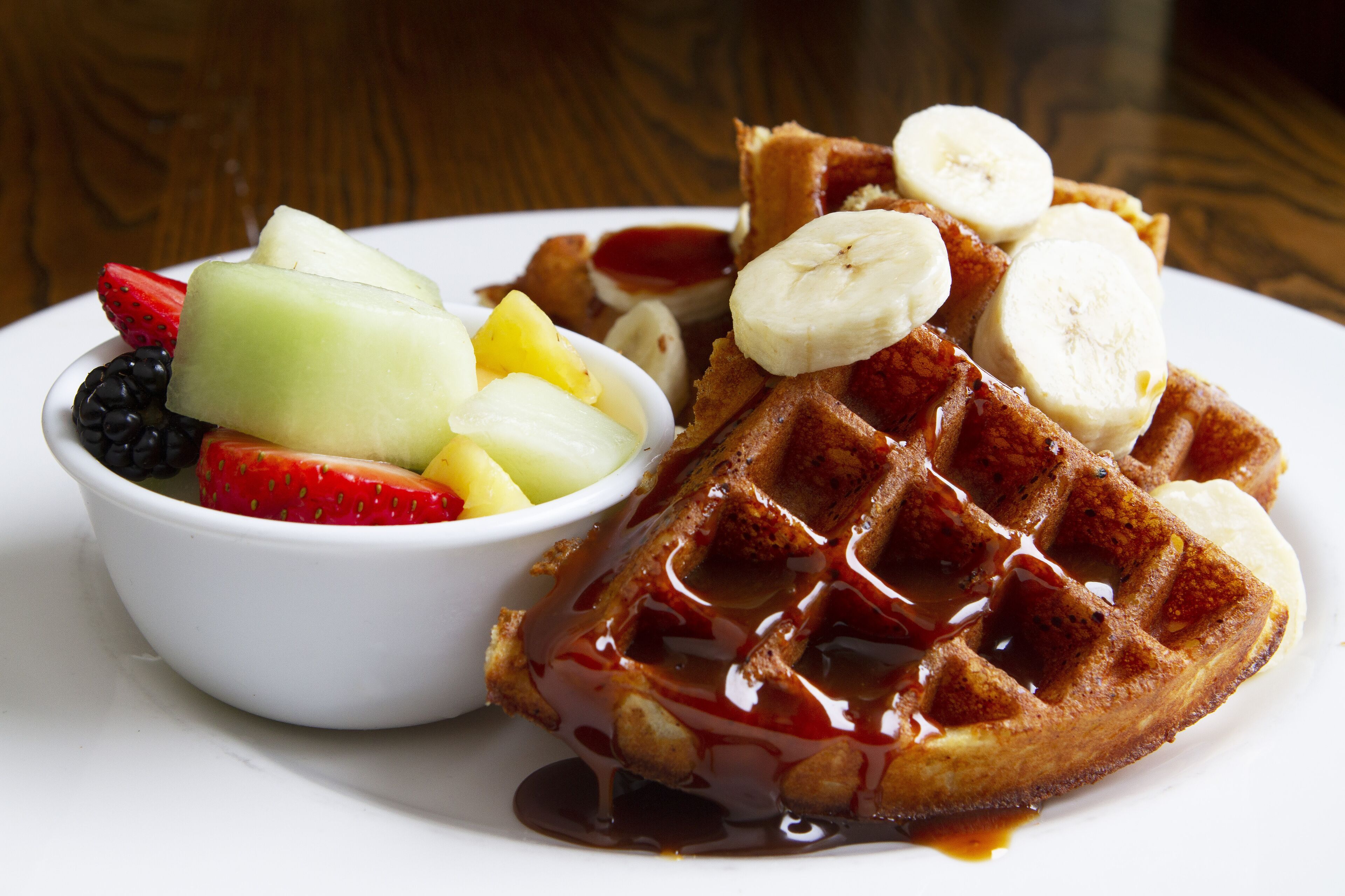 A plate with waffles, syrup, and a bowl of fruit.