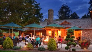 An outdoor patio area with tables and chairs, surrounded by greenery and a stone building with a chimney. - Chetola Resort (Blowing Rock)