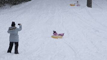 Paisaje nevado con una persona fotografiando, un niño esquiando en trineo y un muñeco de nieve.