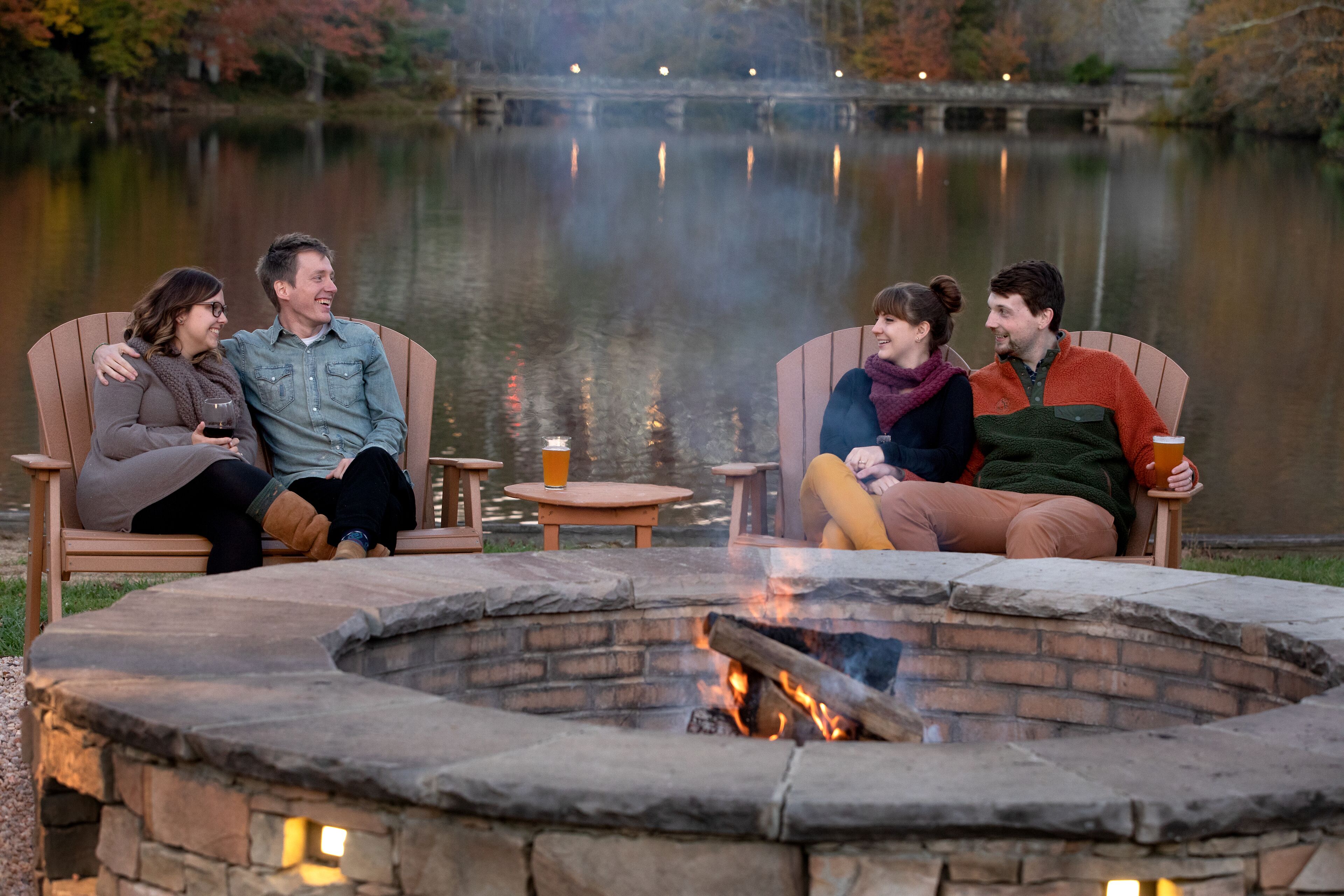 A group of people sitting around a stone fire pit with a lake in the background.