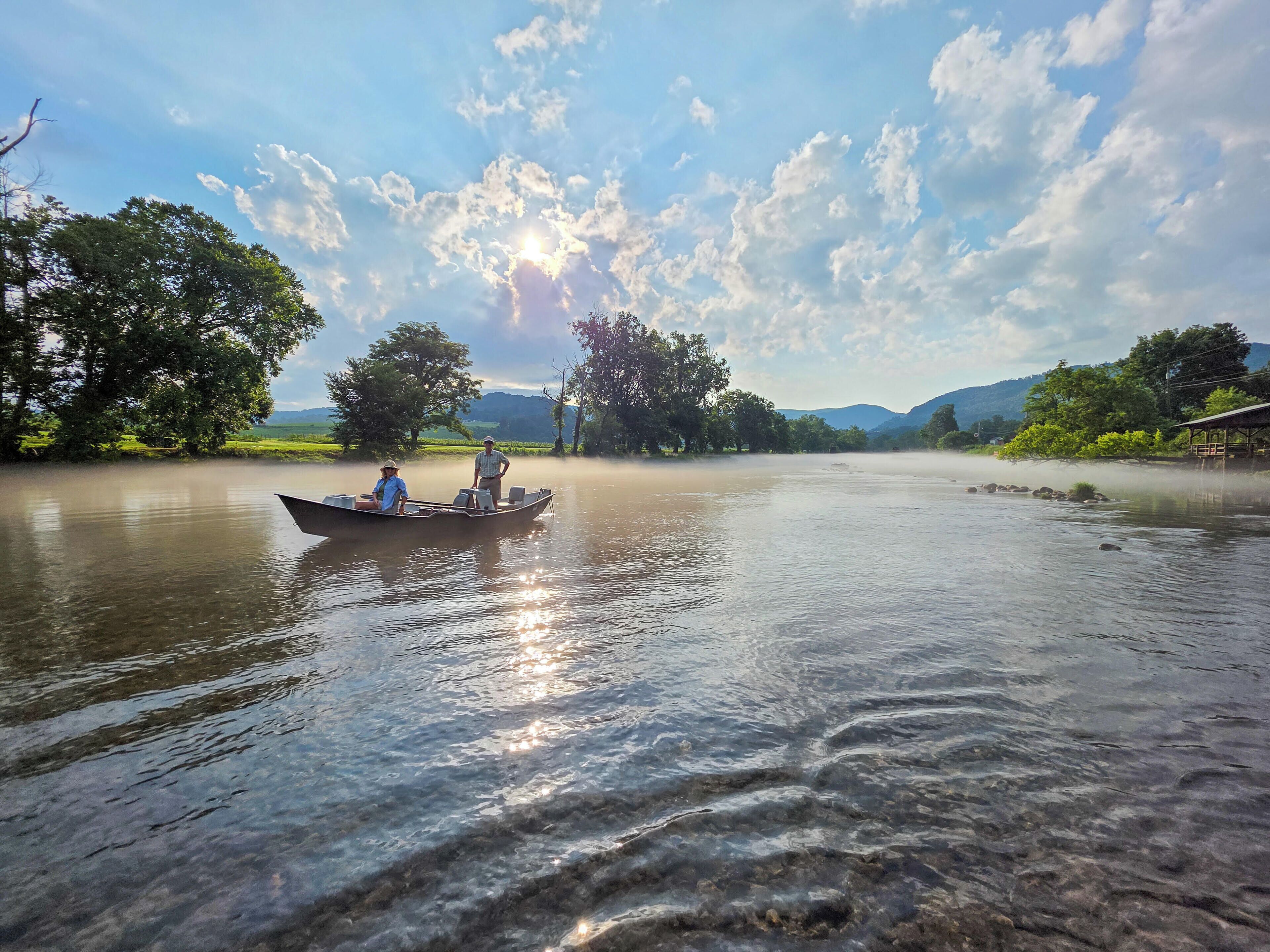 Ein ruhiger Fluss mit einem Boot, das zwei Personen befördert, umgeben von üppiger Vegetation und wolkenlosem Himmel.