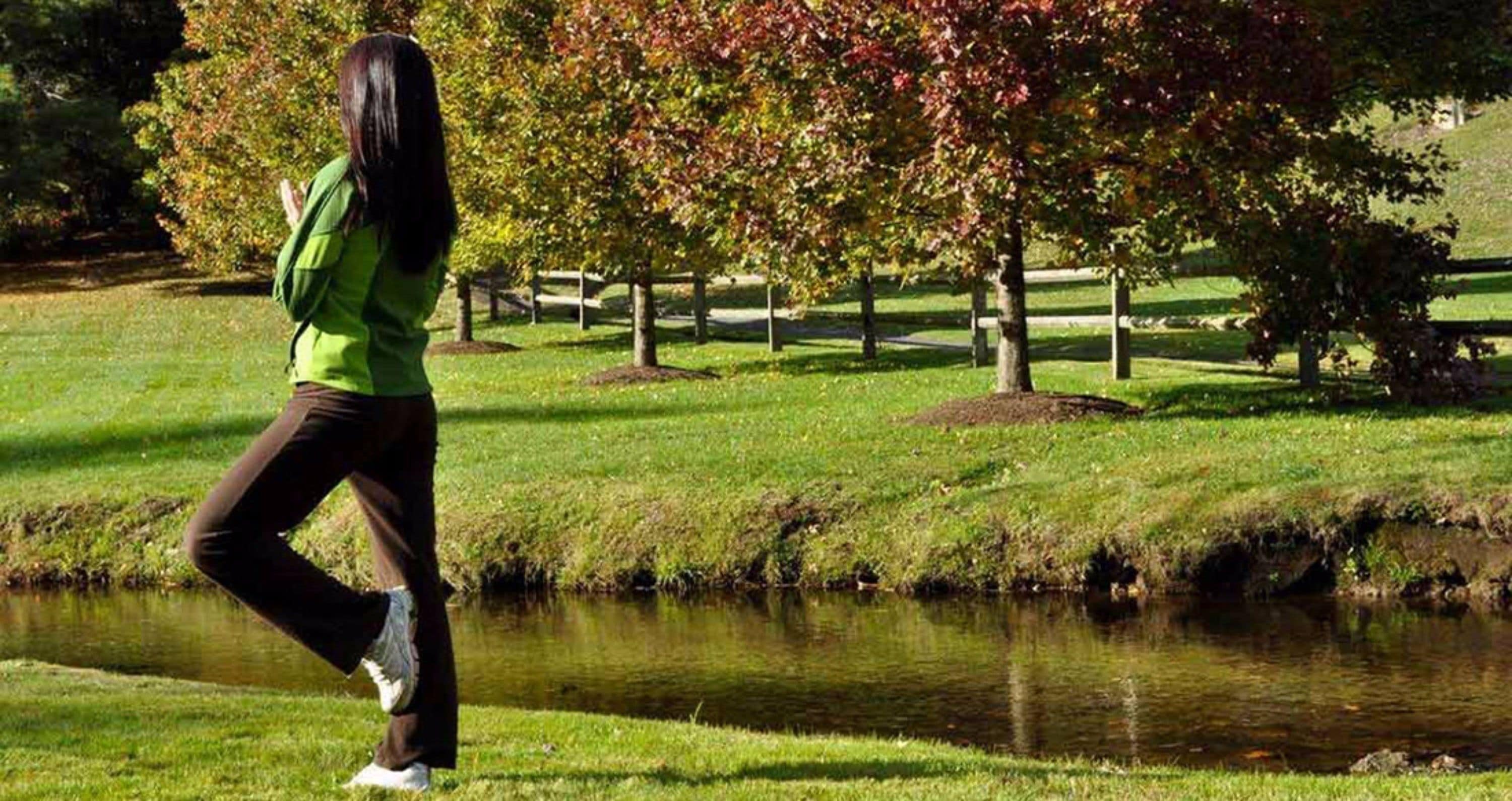 A person walking on a grassy area with a pond and trees in the background.