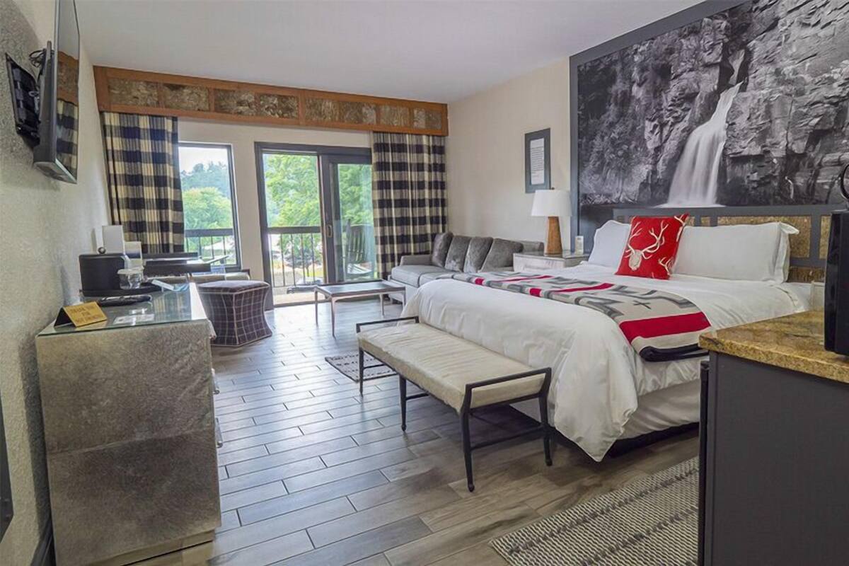 Une chambre d’hôtel avec un lit, un bureau, une banquette, une télévision et une grande fenêtre avec des rideaux.