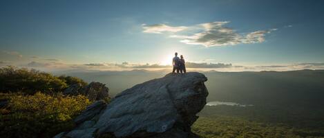 Una pareja de pie sobre una roca, con vista a un valle que incluye un lago y montañas a lo lejos.