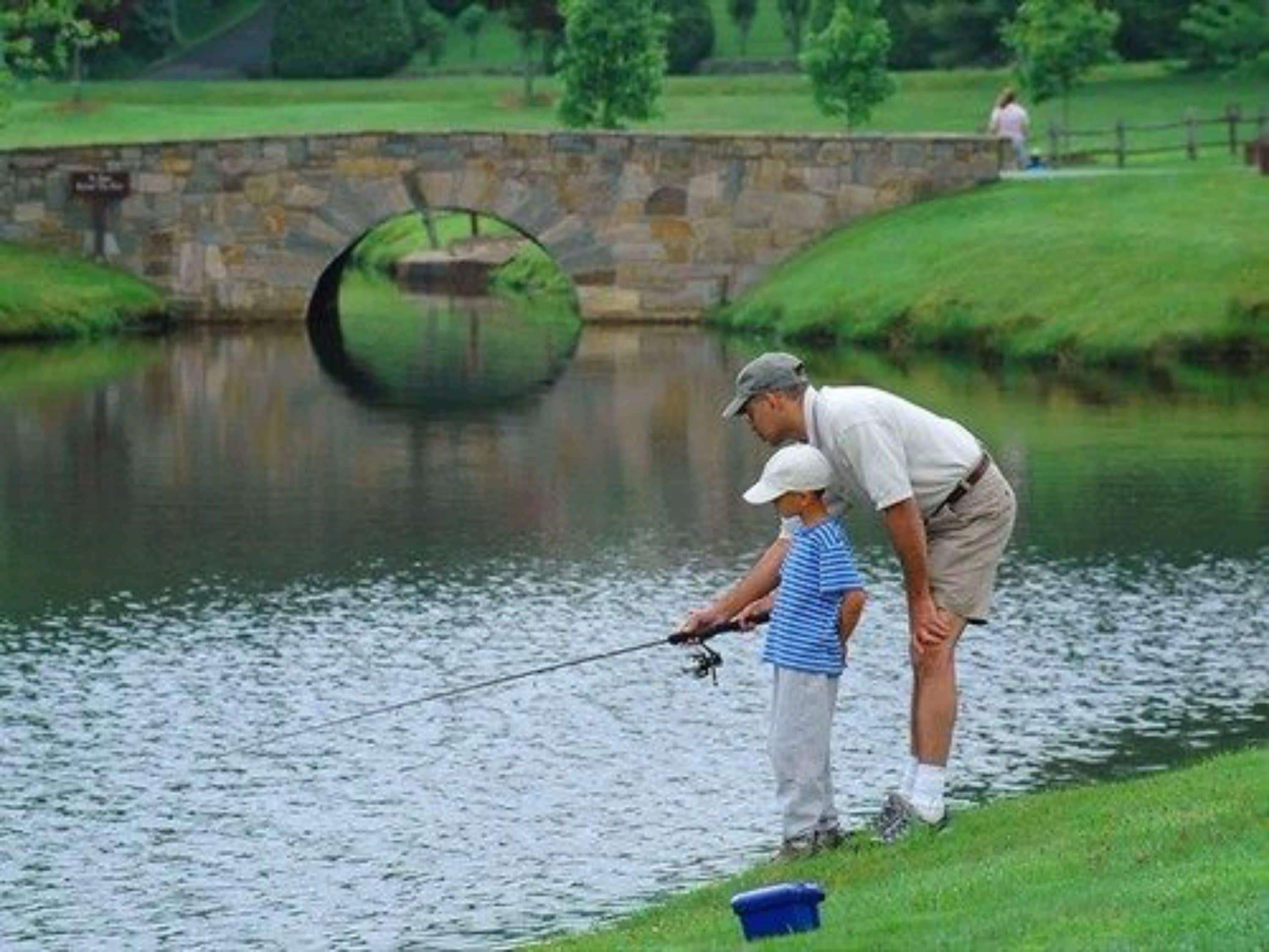 A man and a child fishing by a calm river with a stone bridge in the background.
