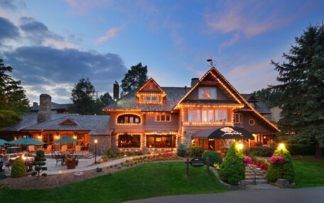 A large, two-story house with a well-lit exterior, a stone chimney, and a landscaped front yard with a pathway leading to the entrance.
