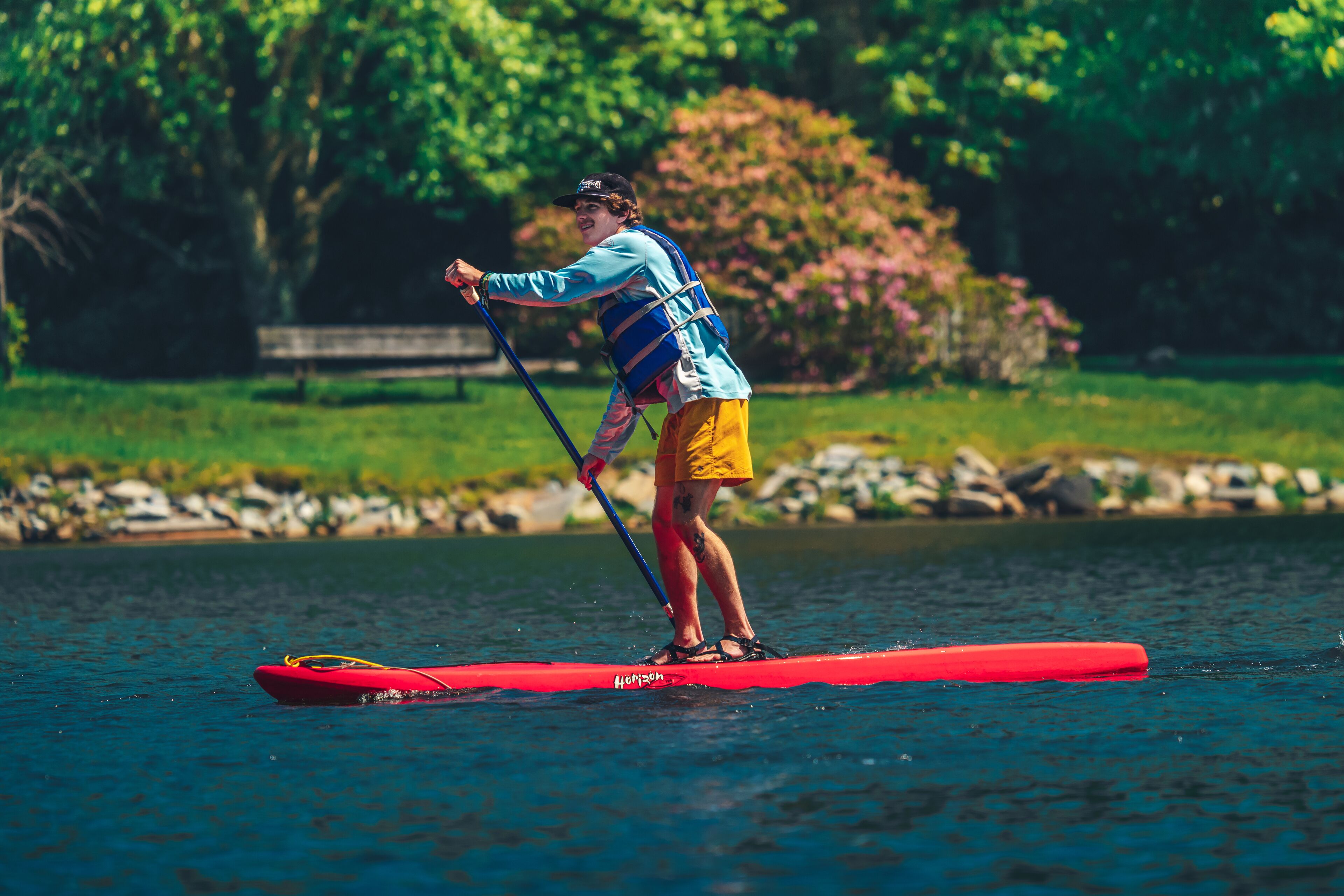 A person standing on a red paddleboard in a body of water, surrounded by greenery and a stone wall.