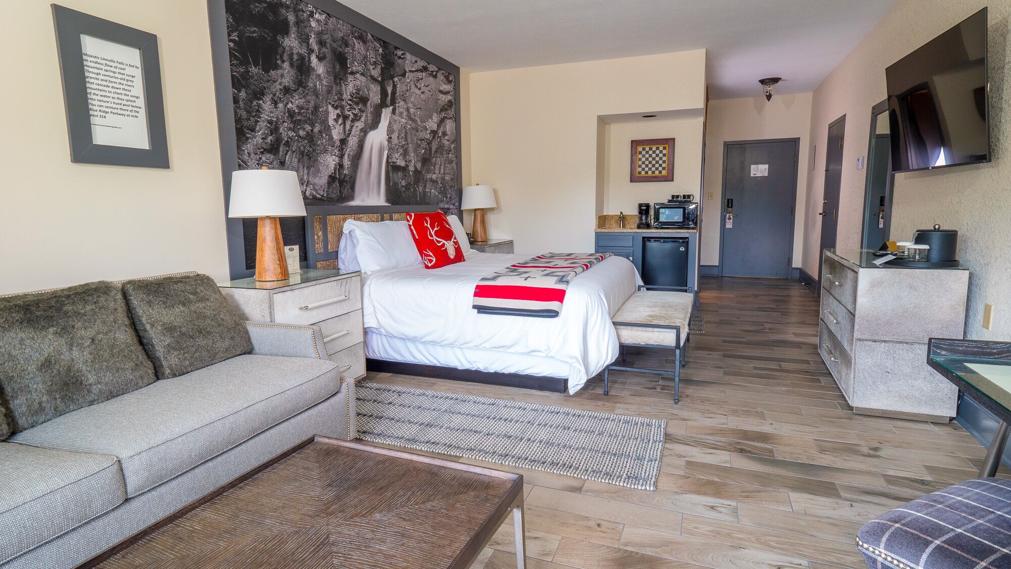 A hotel room with a bed, a sofa, a small table, a desk, a TV, and a kitchenette.