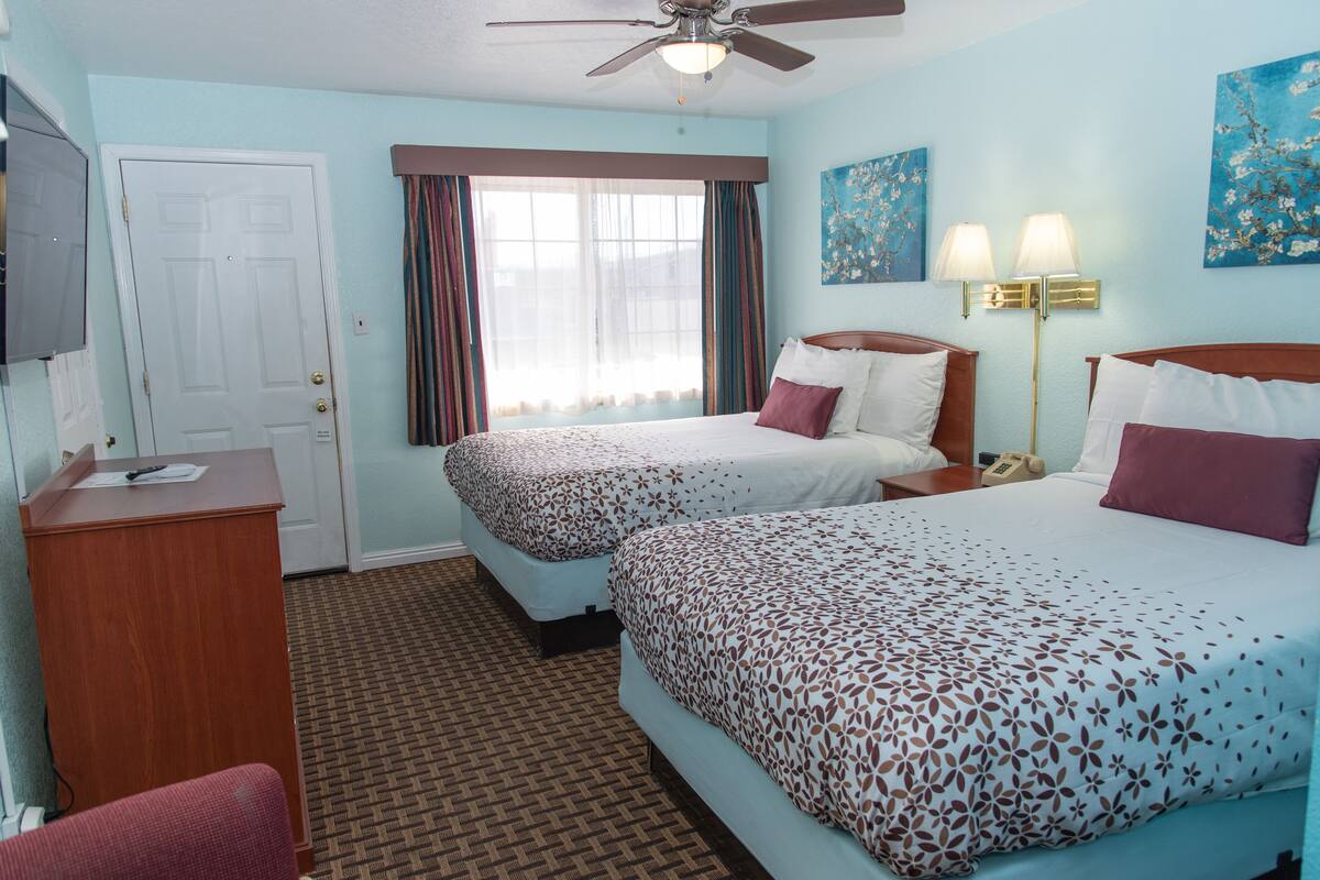 A hotel room with two beds, a desk, a ceiling fan, a window with curtains, and artwork on the walls.