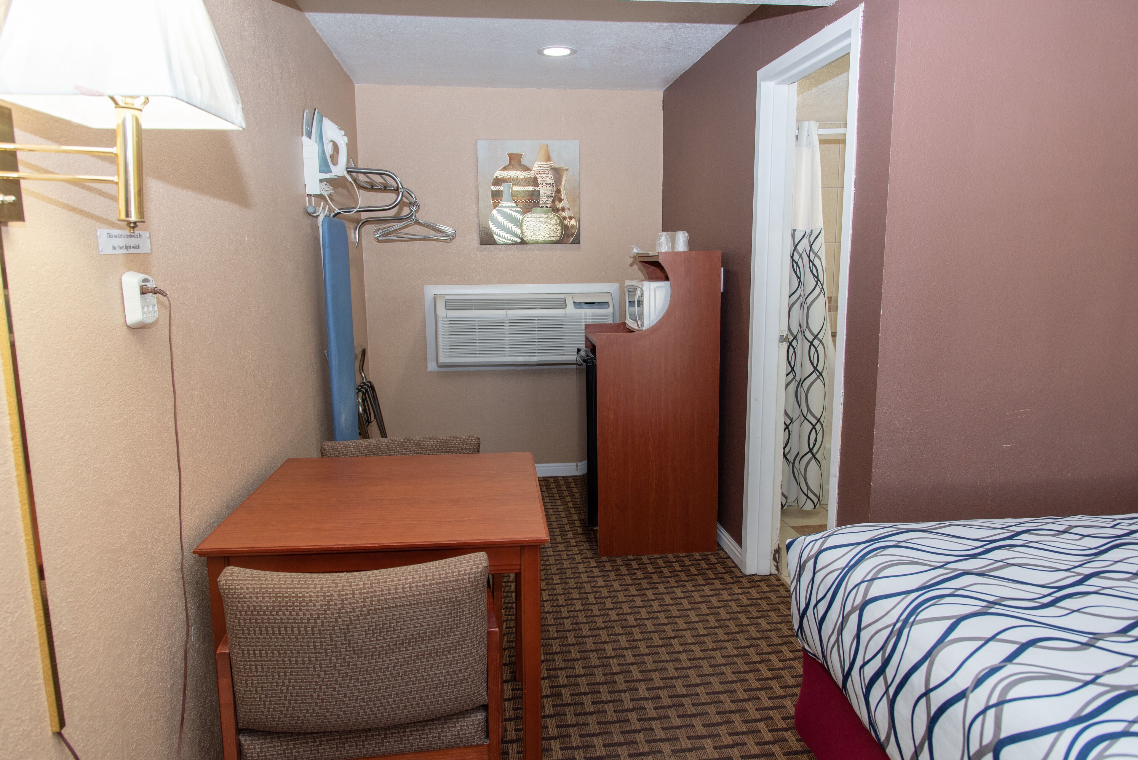 A hotel room with a bed, a desk, a chair, an air conditioning unit, and a wall-mounted lamp.
