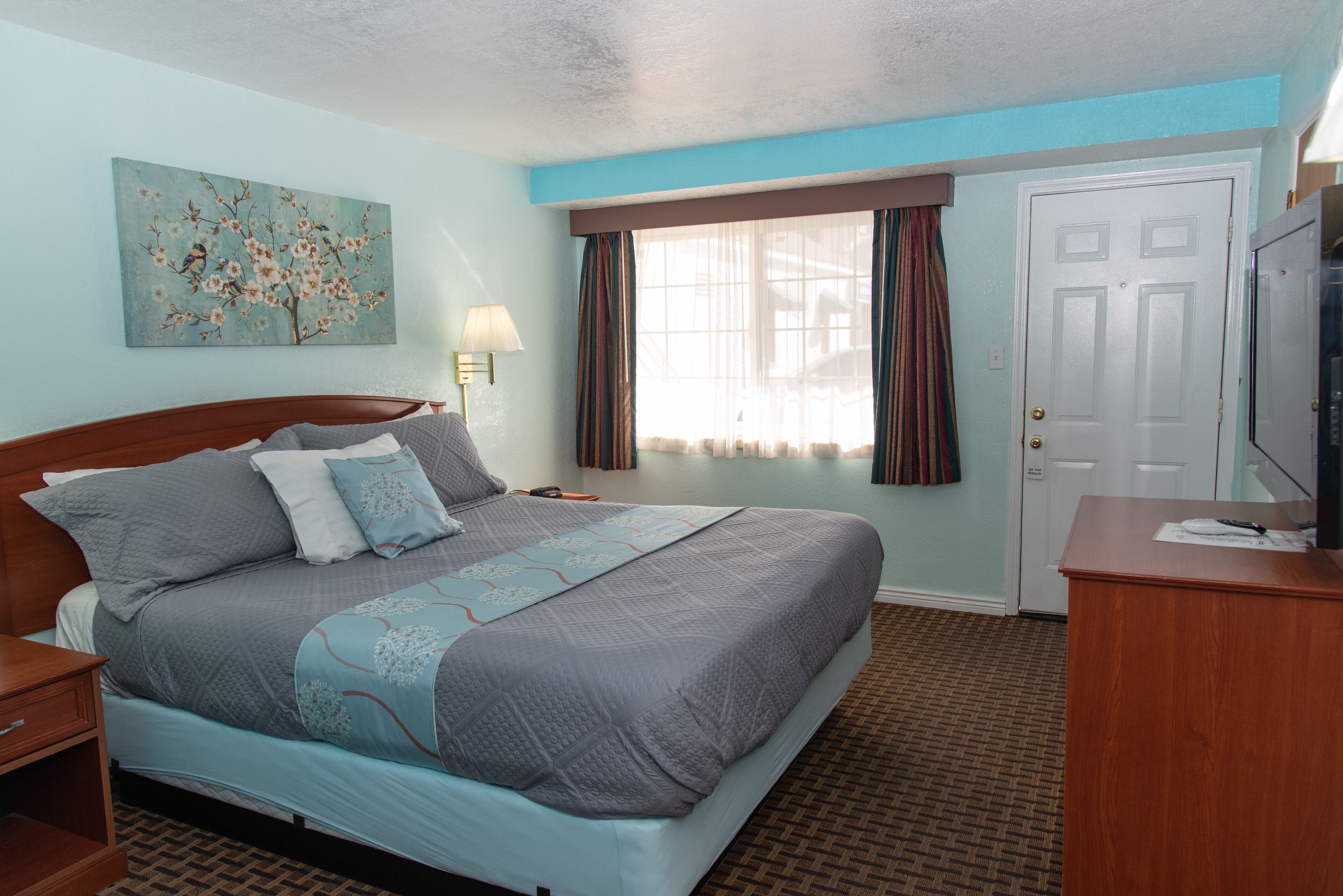 A neatly made bed with a grey and blue quilt, a wooden nightstand, a lamp, a window with curtains, and a door.
