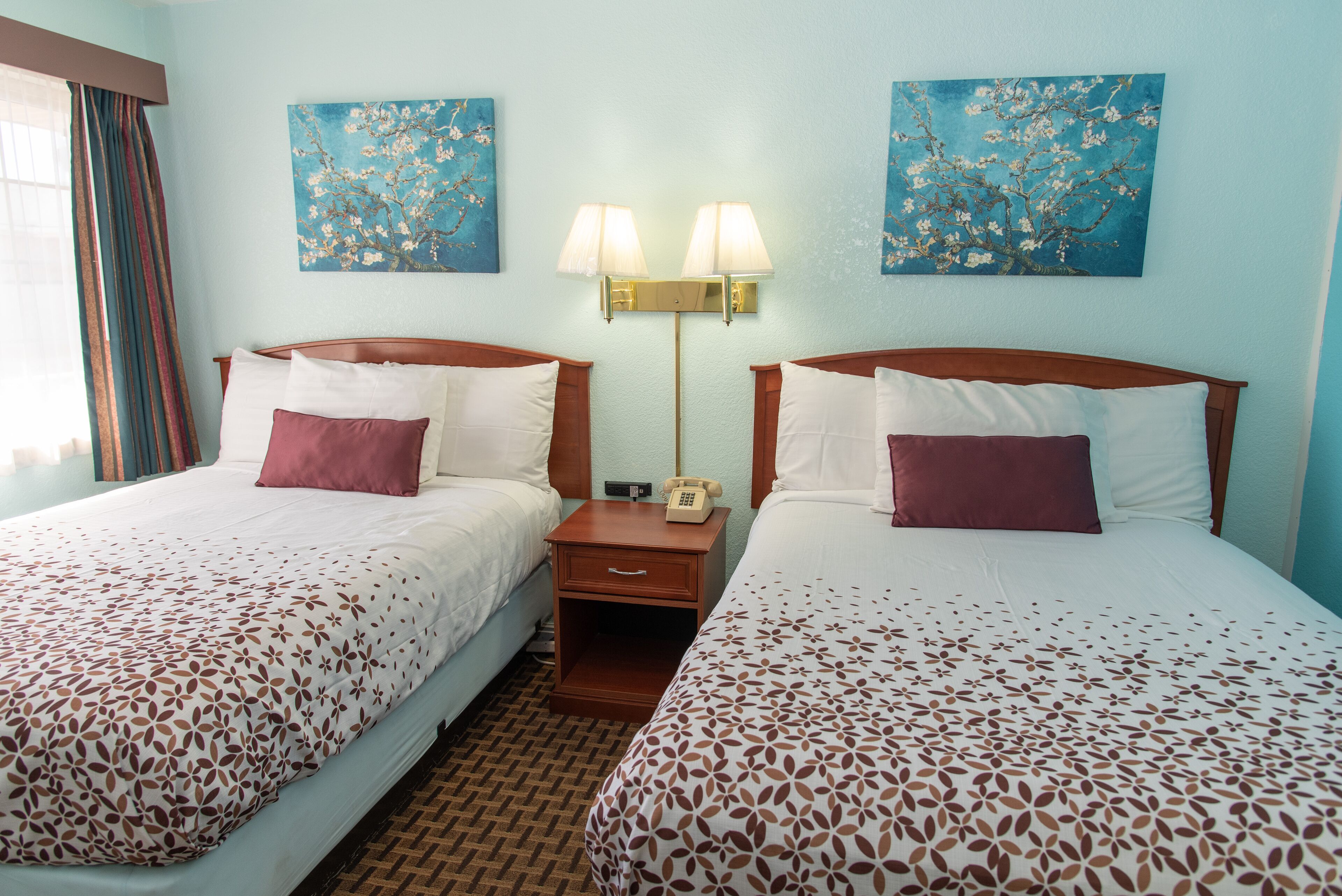 A hotel room with two beds, each with a patterned bedspread, a nightstand with a lamp, and two wall-mounted paintings.