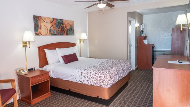 A hotel room with a bed, bedside tables, a desk, a chair, a ceiling fan, and a painting on the wall.