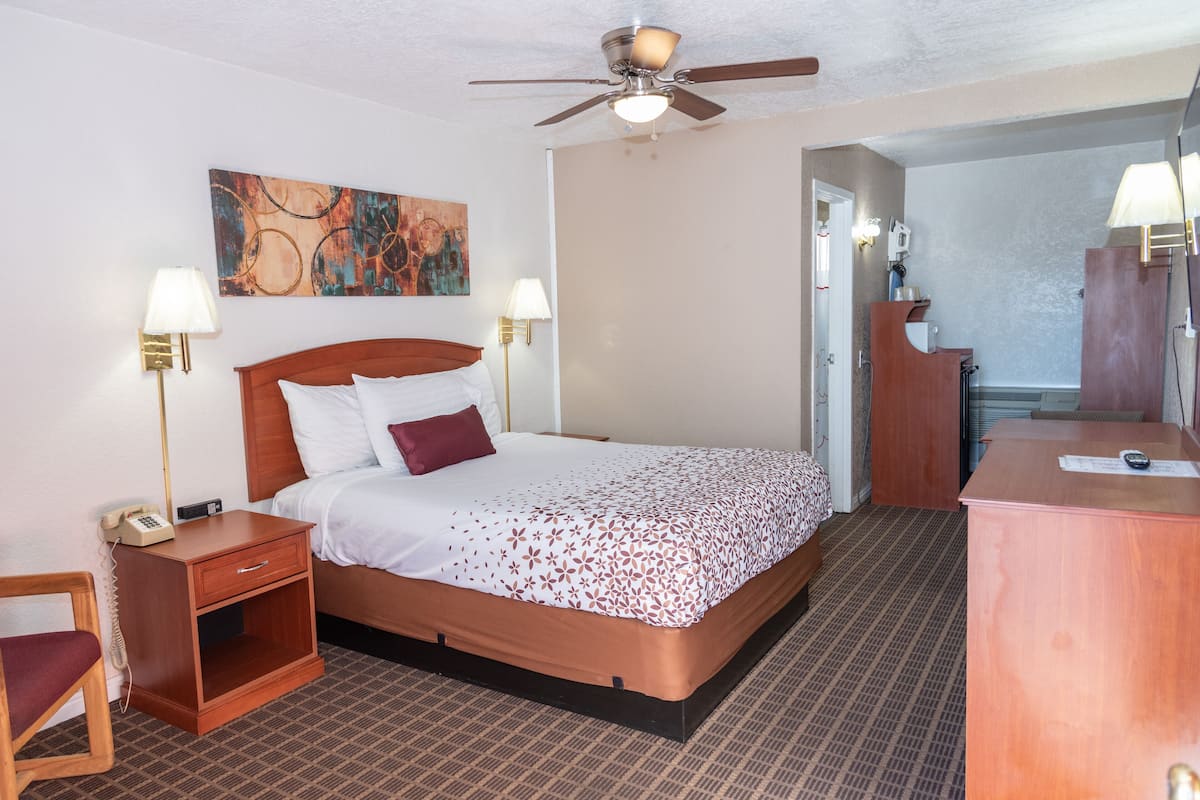 A hotel room with a bed, bedside tables, a desk, a chair, a ceiling fan, and a painting on the wall.