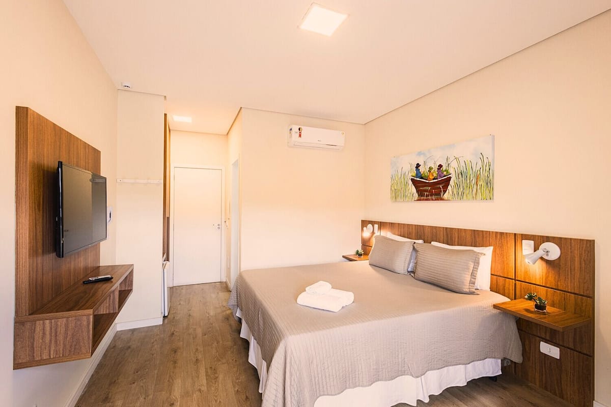 A bedroom with a bed, a TV mounted on a wooden panel, a painting on the wall, and a door leading to another room.