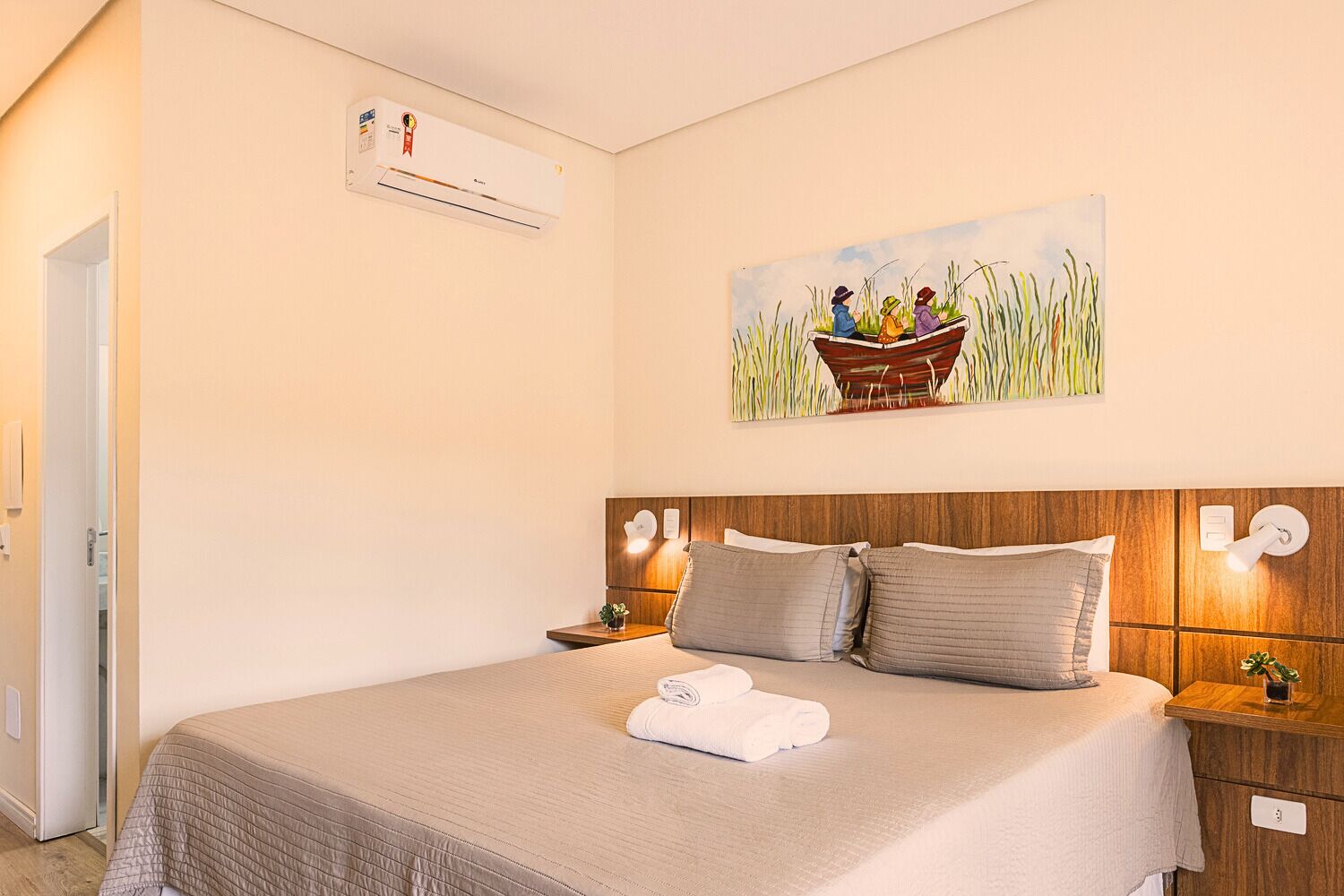 A neatly arranged hotel room with a bed, a painting of a boat on a river, and a wall-mounted air conditioner.