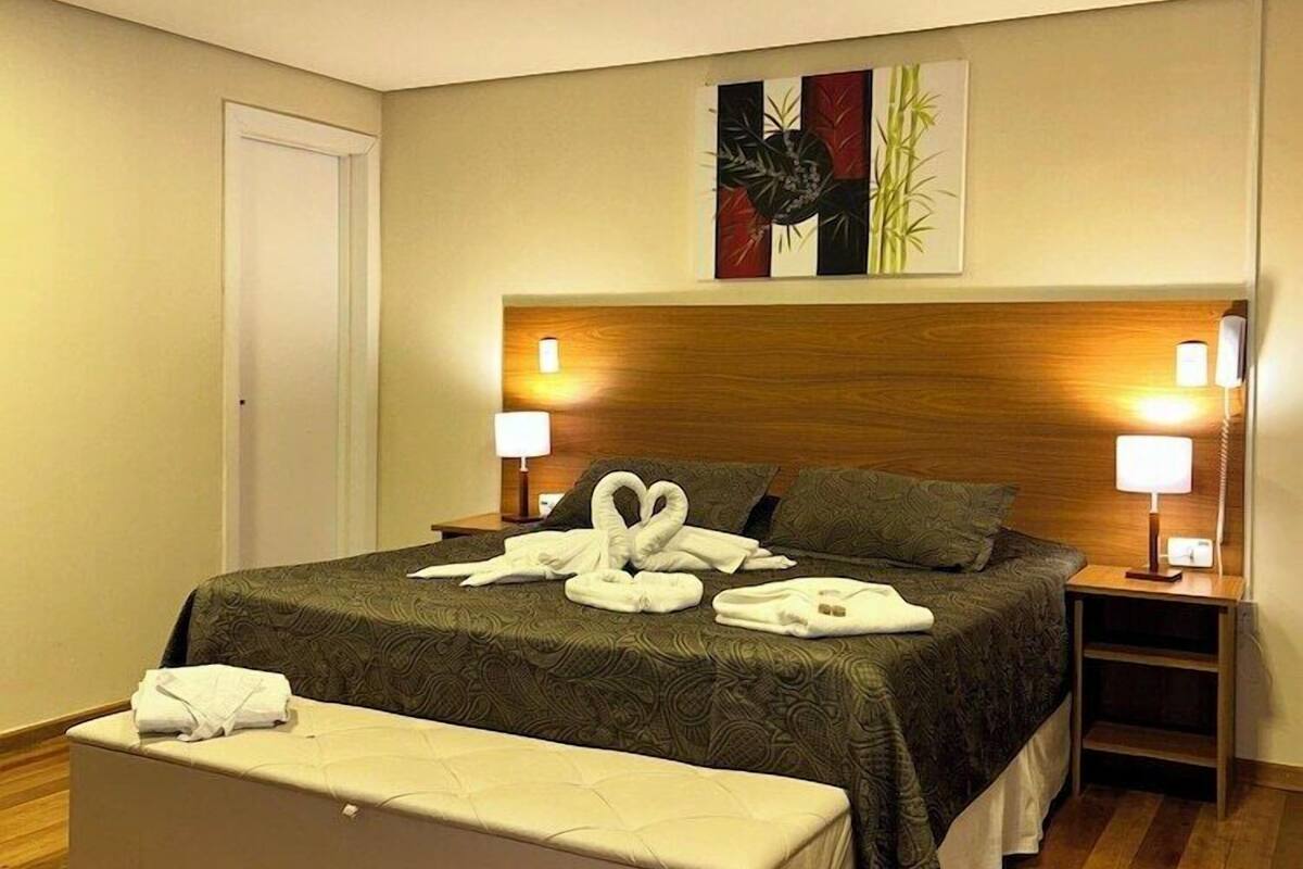 A hotel room with a bed, bedside lamps, a bench, and a painting on the wall.