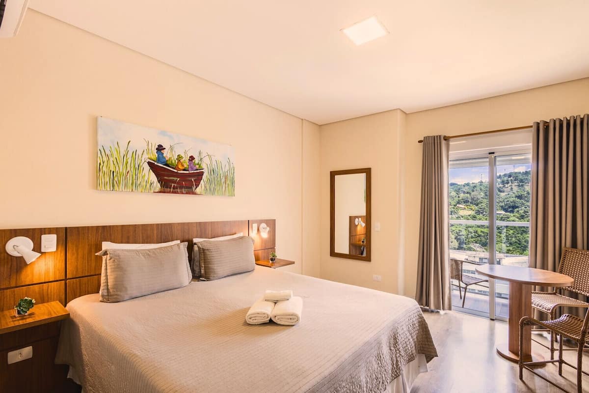 A hotel room with a large bed, a painting of people in a boat, a balcony with a table and chairs, and a view of greenery.