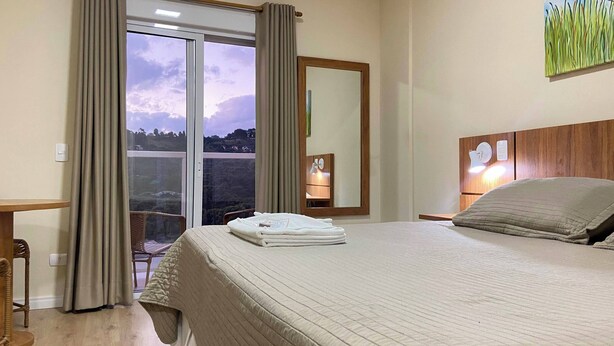 A hotel room with a bed, a nightstand, a mirror, and a view of the outdoors. - Kaliman Campos do Jordão (Campos do Jordão)
