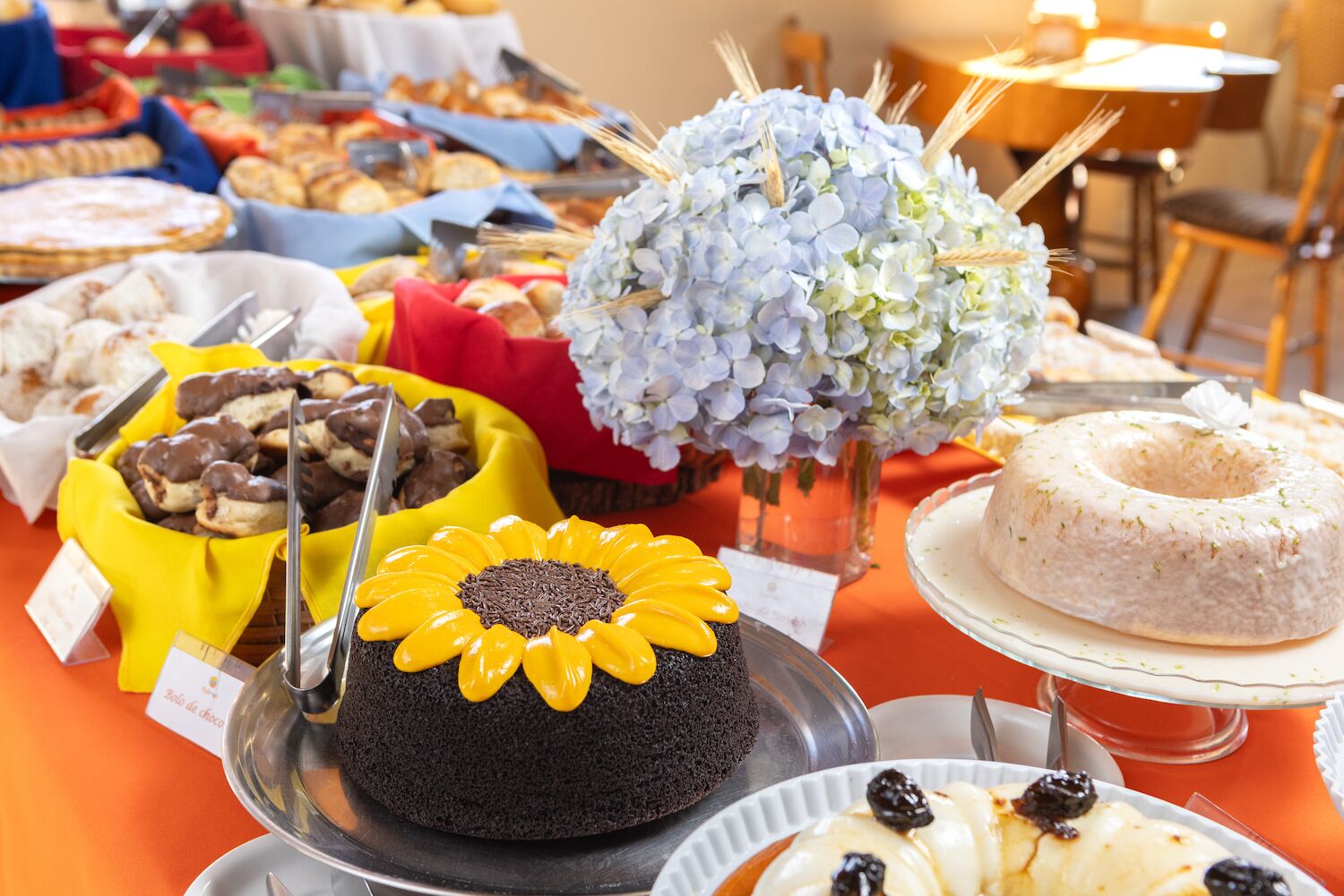 Un tavolo del buffet con diversi cibi, tra cui una torta al cioccolato decorata con un fiore giallo, una torta bianca con una coroncina e un'ampia composizione floreale.