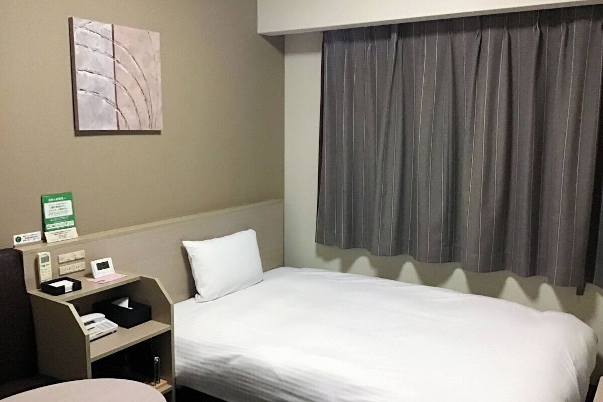 A hotel room with a single bed, a bedside table with a telephone and a small box, a pillow, a wall-mounted artwork, and a window with curtains.