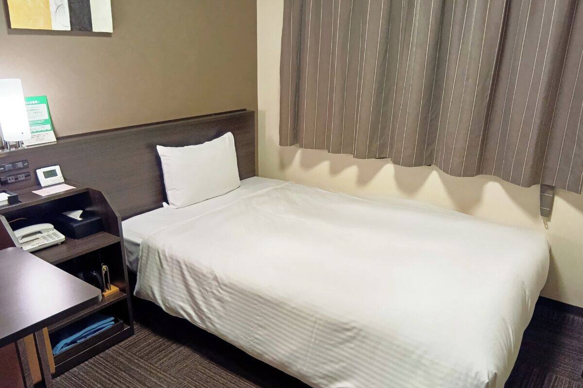 A hotel room with a single bed, a bedside table with a phone and a lamp, and a small shelf with a tissue box and a book.