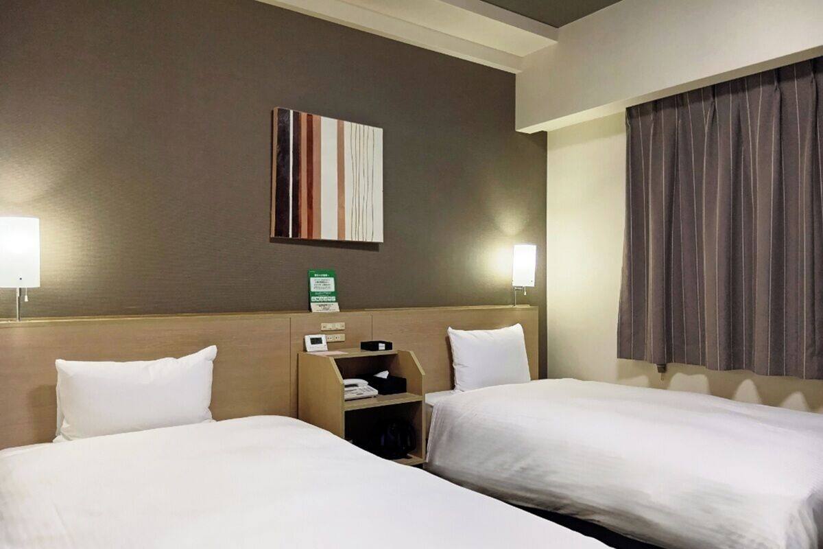 A hotel room with two beds, a bedside table with a phone and a small shelf, a wall-mounted artwork, and a window with curtains.