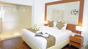 A hotel room with a large bed, a nightstand with a lamp, a decorative headboard, and a shower area. - Samsara Inn (Kuta)