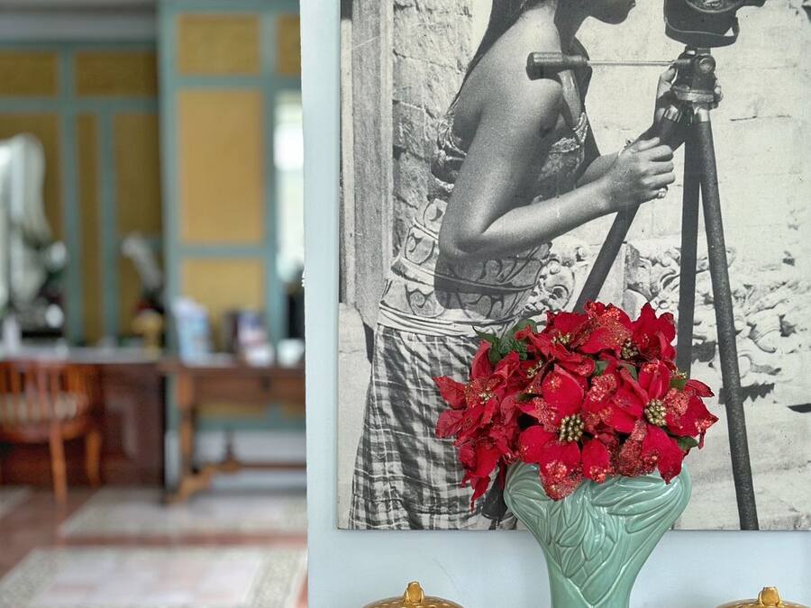 Une photographie représentant une femme tenant un appareil photo sur un trépied, un vase de fleurs rouges et deux petits pots jaunes posés sur une table en bois.