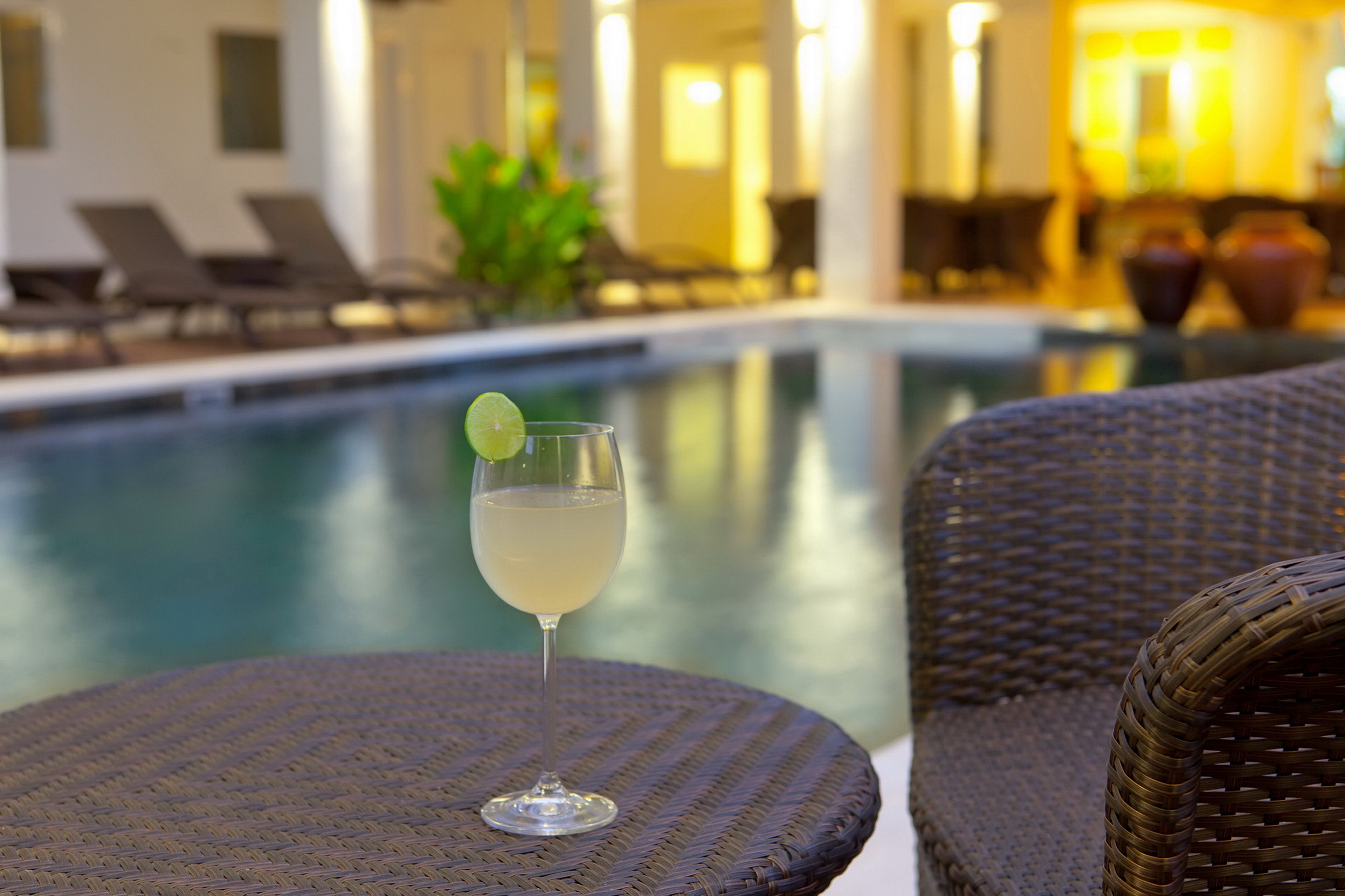 A poolside setting with a glass of a light-colored drink garnished with a lime, wicker furniture, and a pool in the background.